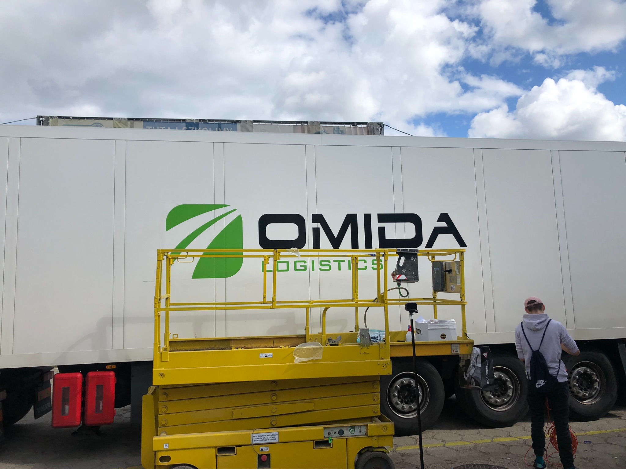 Pracownik na podnośniku koszowym okleja naczepę ciężarówki logo firmy Omida Logistics, widoczne narzędzia i materiały przygotowane do pracy.