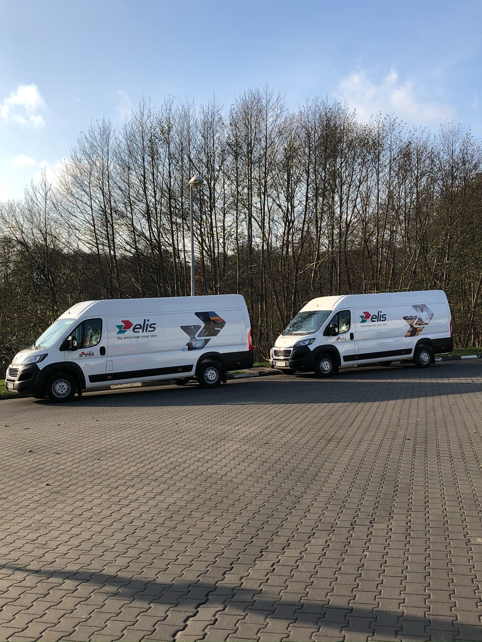 Dwa białe samochody dostawcze z logo firmy Elis zaparkowane na brukowanym parkingu, z drzewami bez liści w tle i błękitnym niebem.