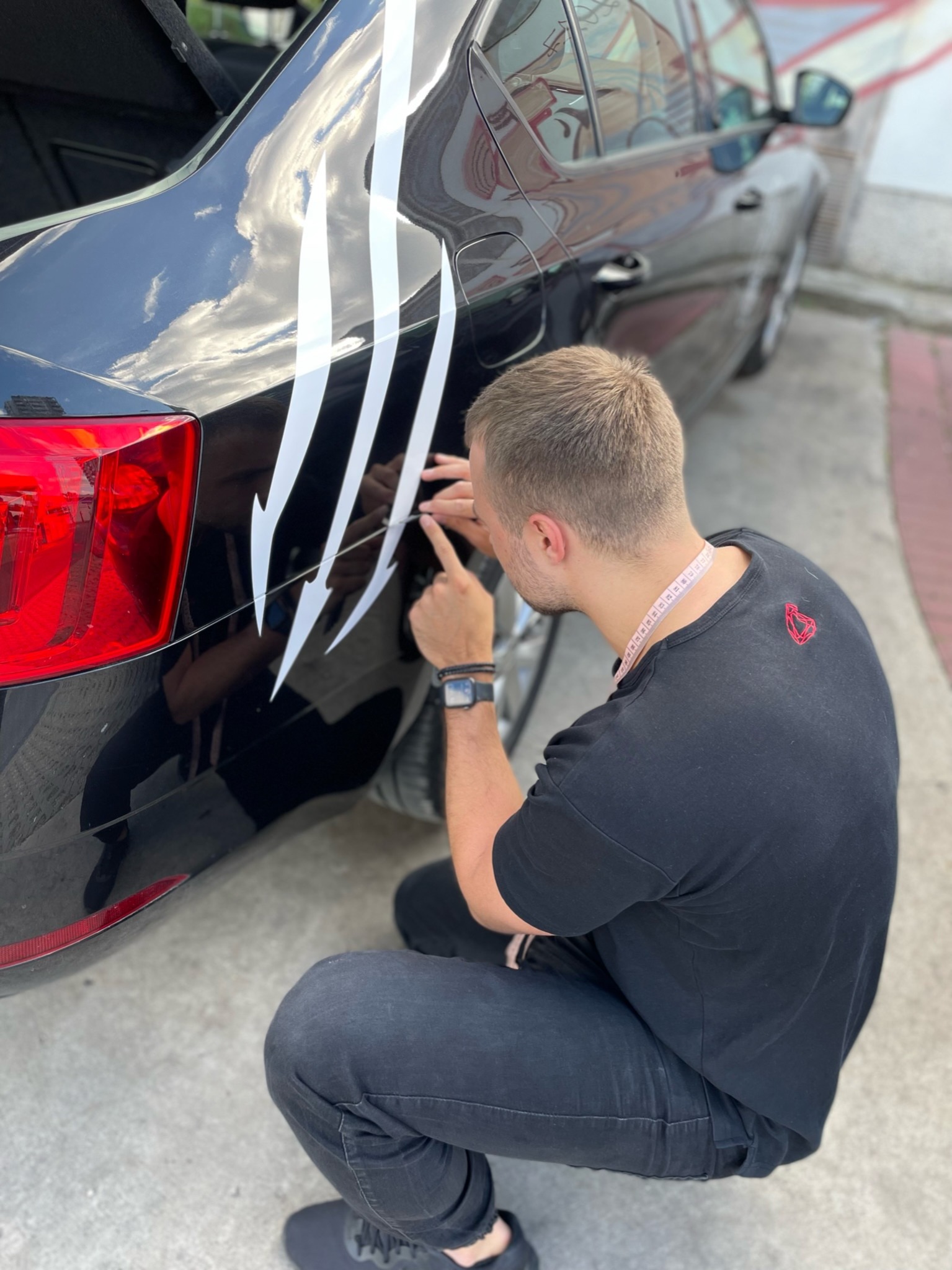 Oklejanie czarnego samochodu naklejkami imitującymi ślady pazurów; pracownik precyzyjnie nakleja białą folię na karoserię, używając narzędzia do aplikacji.