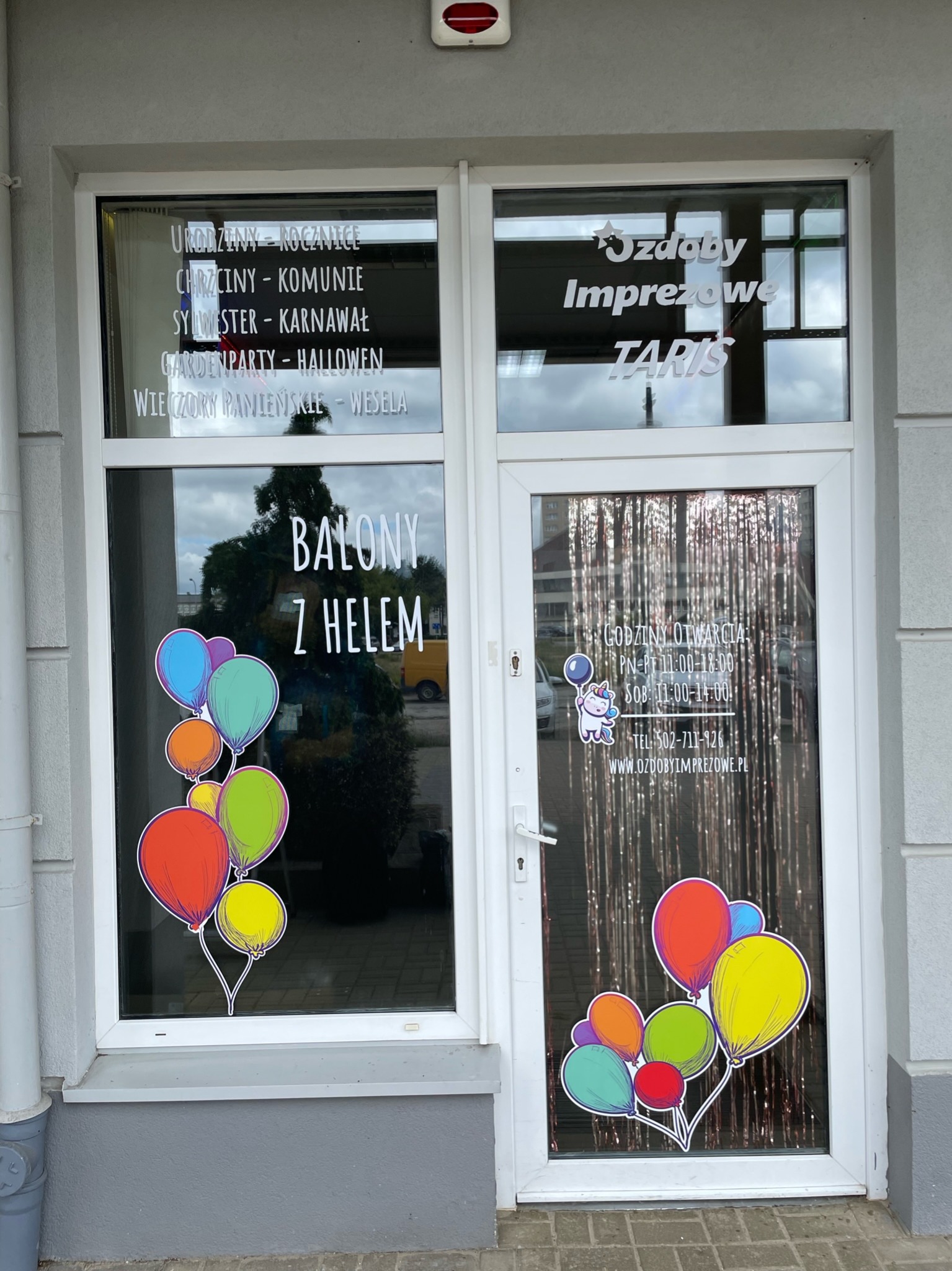 Okna i drzwi lokalu z naklejonymi informacjami o ofercie: balony z helem, dekoracje na imprezy okolicznościowe, godziny otwarcia i kontakt. Na szybach naklejone kolorowe rysunki balonów.