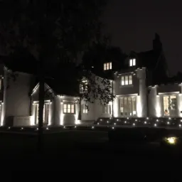 Nowoczesny, biały dom jednorodzinny oświetlony zewnętrznymi lampami LED wzdłuż elewacji i schodów wejściowych, widoczny nocą, z ciemnym niebem i drzewami w tle.