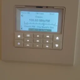 Panel sterowania inteligentnego domu z wyświetlaczem tunera FM, pokazujący stację Classic FM na częstotliwości 100.60 MHz, z przyciskami dotykowymi do nawigacji i regulacji głośności.