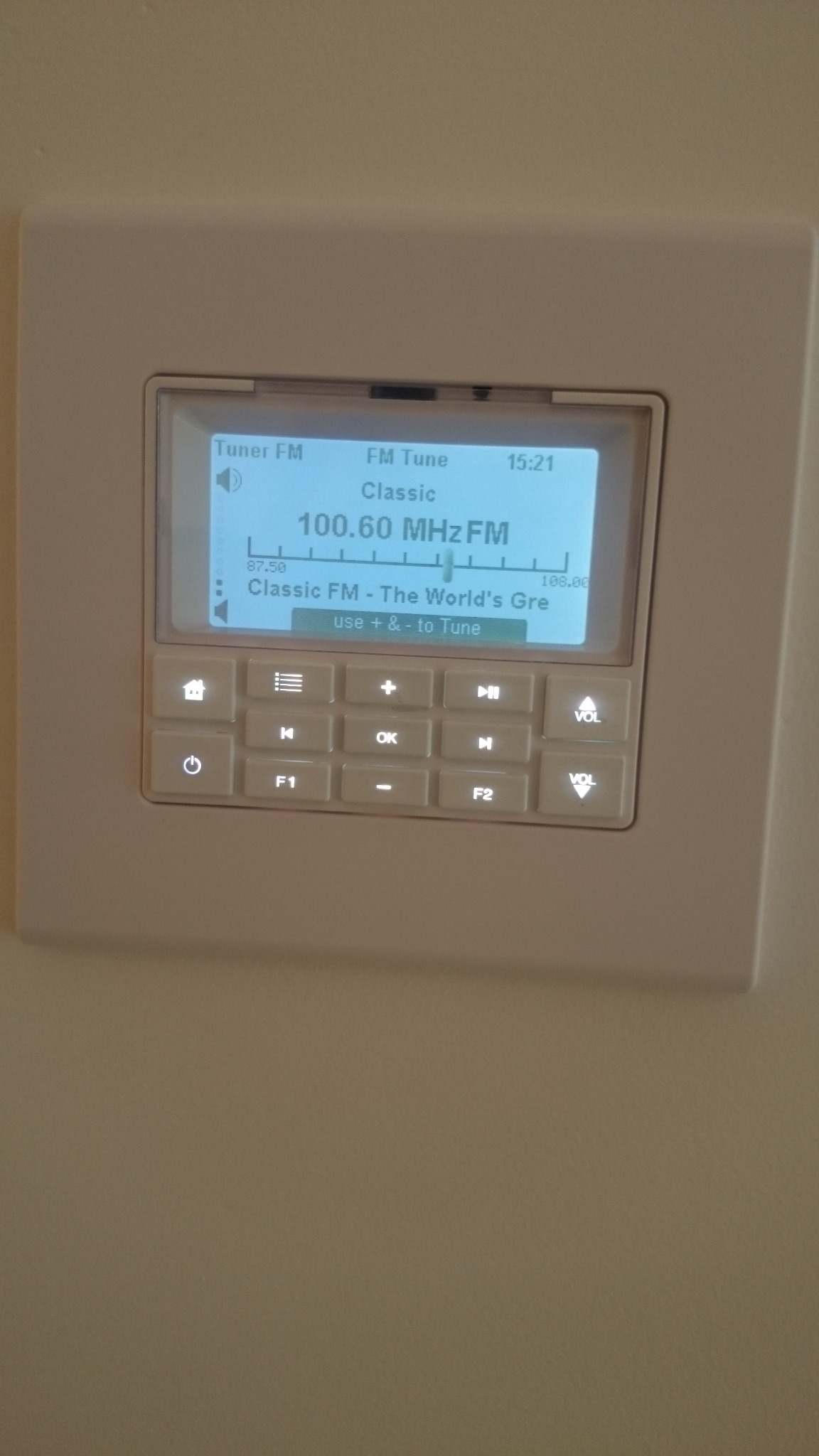 Panel sterowania inteligentnego domu z wyświetlaczem tunera FM, pokazujący stację Classic FM na częstotliwości 100.60 MHz, z przyciskami dotykowymi do nawigacji i regulacji głośności.