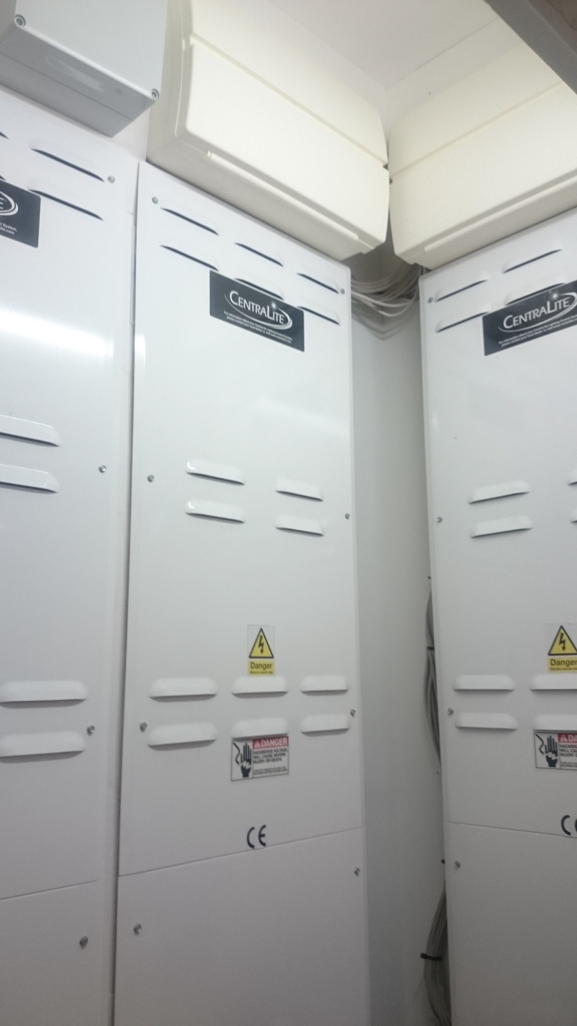 Dwa białe panele sterowania Centralite zamontowane obok siebie w rogu pomieszczenia, z widocznymi przewodami u góry i naklejkami ostrzegawczymi o niebezpieczeństwie porażenia prądem.