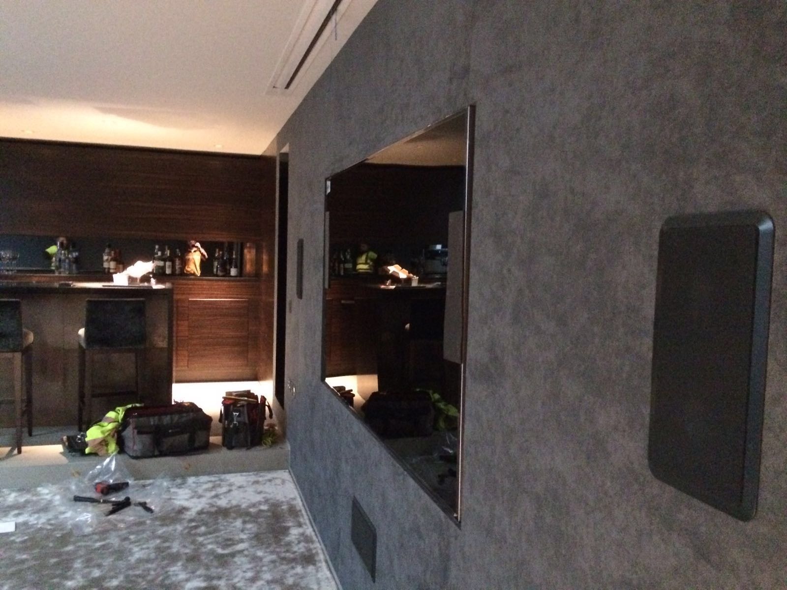 Instalacja głośnika na teksturowanej, szarej ścianie w nowoczesnym wnętrzu z barkiem; widoczne narzędzia i torby instalacyjne na podłodze.