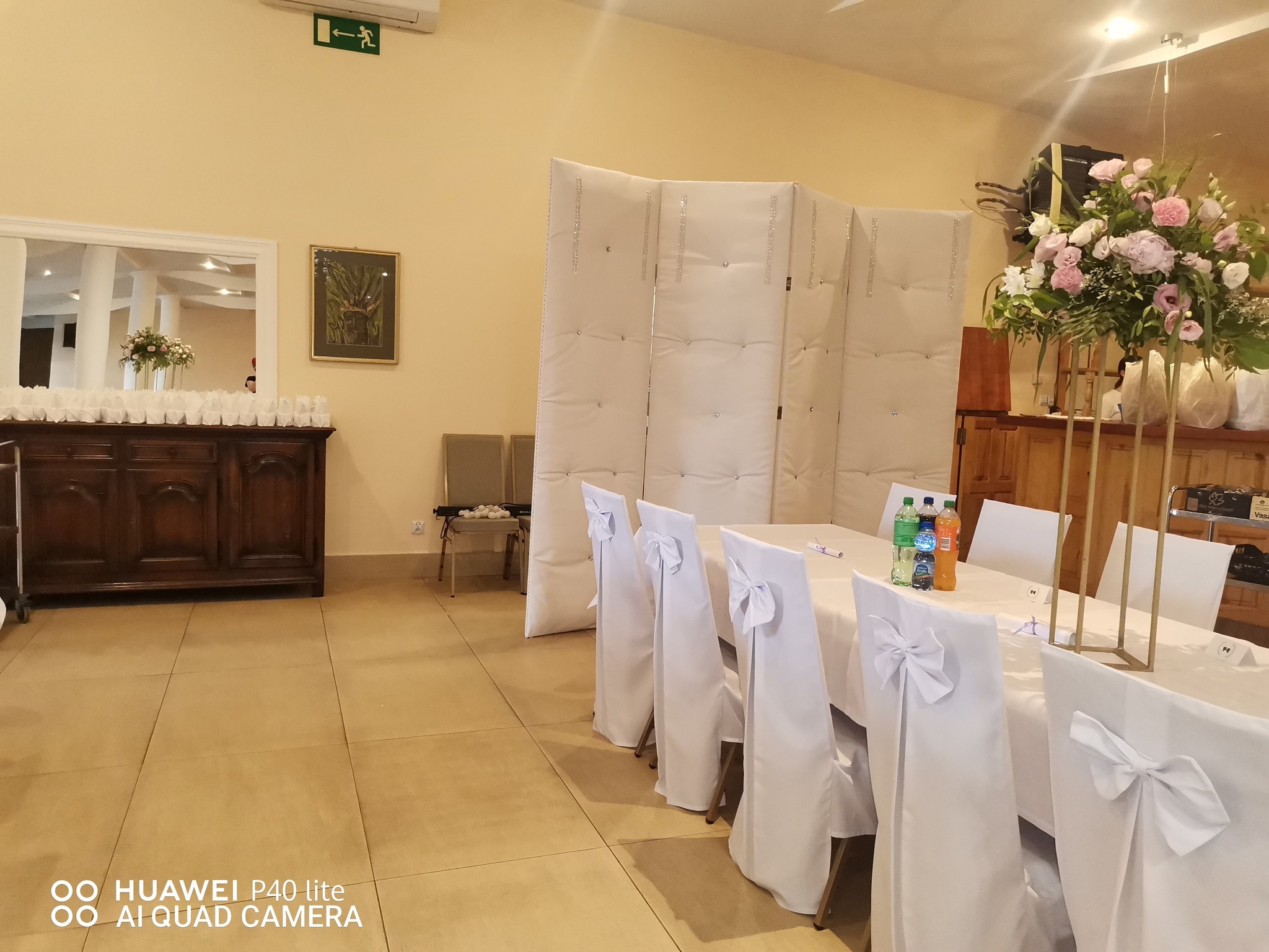 Uroczysta sala z nakrytymi stołami, krzesła z białymi pokrowcami zdobione kokardami, kwiatowe dekoracje na wysokich stojakach, parawan dekoracyjny i ciemna komoda z białymi świecznikami.