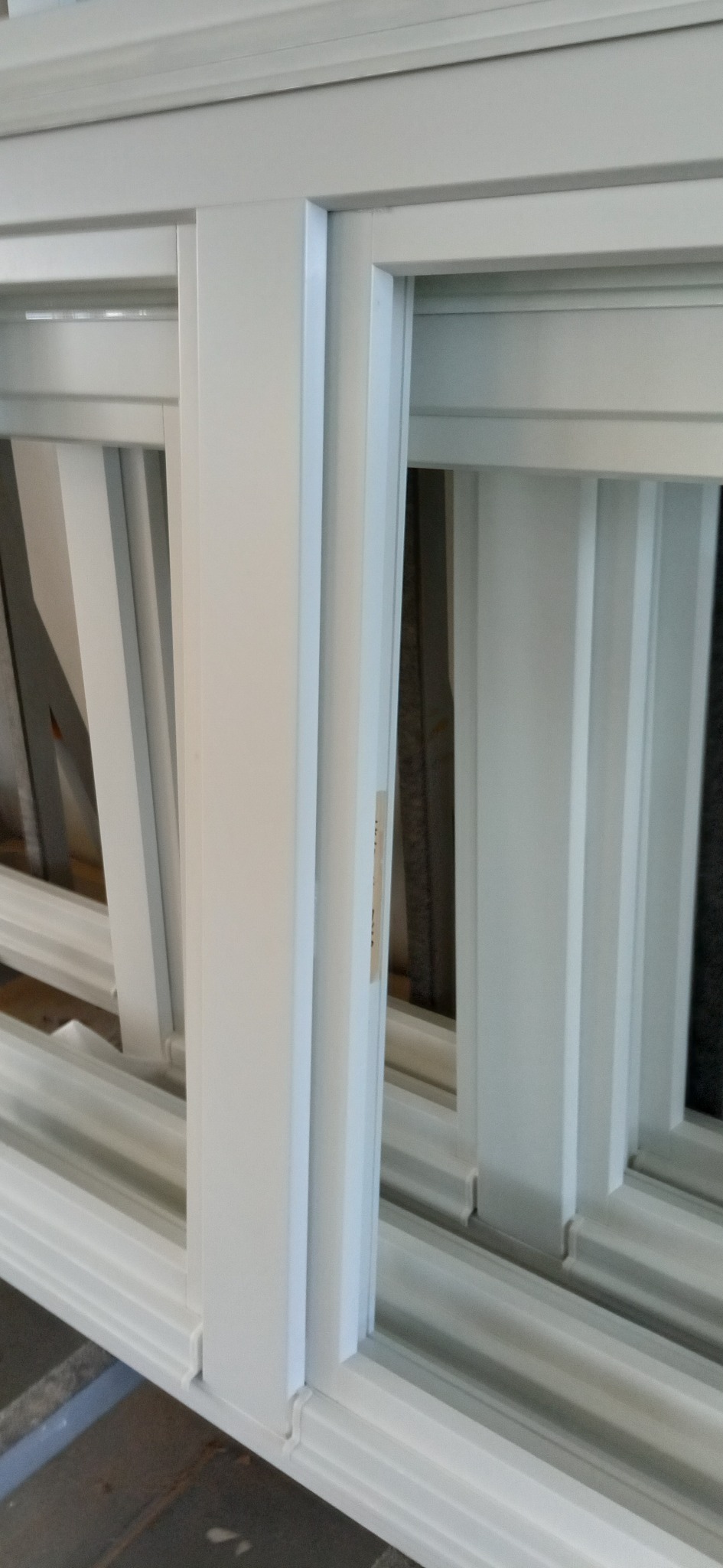 Białe okno przesuwne PCV z widocznymi profilami i prowadnicami, w trakcie montażu lub transportu, z naklejką informacyjną na jednym ze skrzydeł.