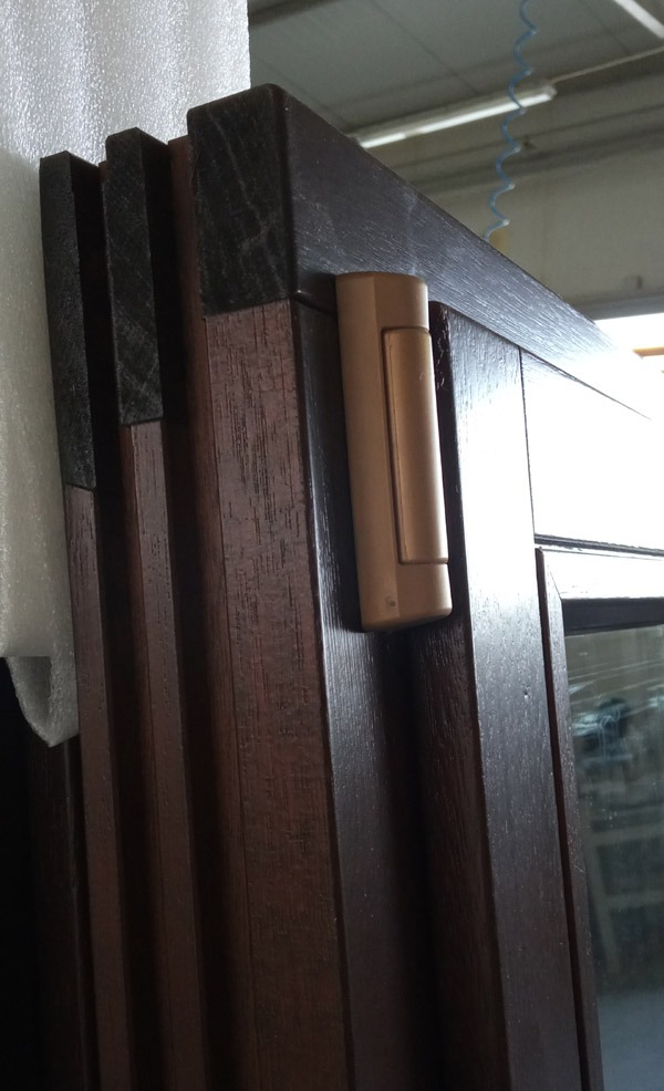 Fragment brązowej ramy okiennej z drewna z widocznym zawiasem, zabezpieczona folią ochronną.