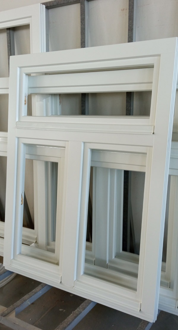 Białe ramy okienne z podwójnymi szybami, ułożone jedna za drugą w magazynie lub warsztacie, widoczne detale wykończenia profili.