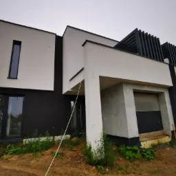 Nowoczesny dom murowany z czarno-białą elewacją, widoczne elementy konstrukcyjne i teren wokół w trakcie prac wykończeniowych, z ziemią i roślinnością.