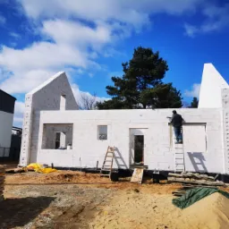 Wznoszenie ścian domu z białych bloczków na tle błękitnego nieba z chmurami, pracownik na drabinie zacierający spoiny, widoczne okna i drzwi w stanie surowym, przygotowany teren budowy z piaskiem...