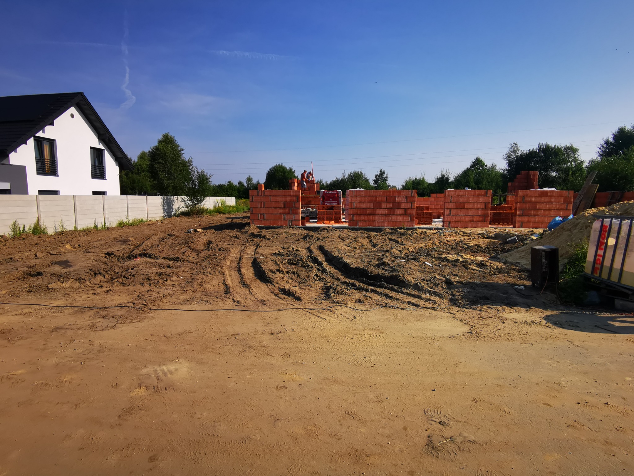Wznoszenie ścian domu z czerwonej cegły ceramicznej Porotherm na placu budowy w słoneczny dzień, widoczni dwaj murarze przy pracy, w tle biały dom z czarnym dachem i zielone drzewa.