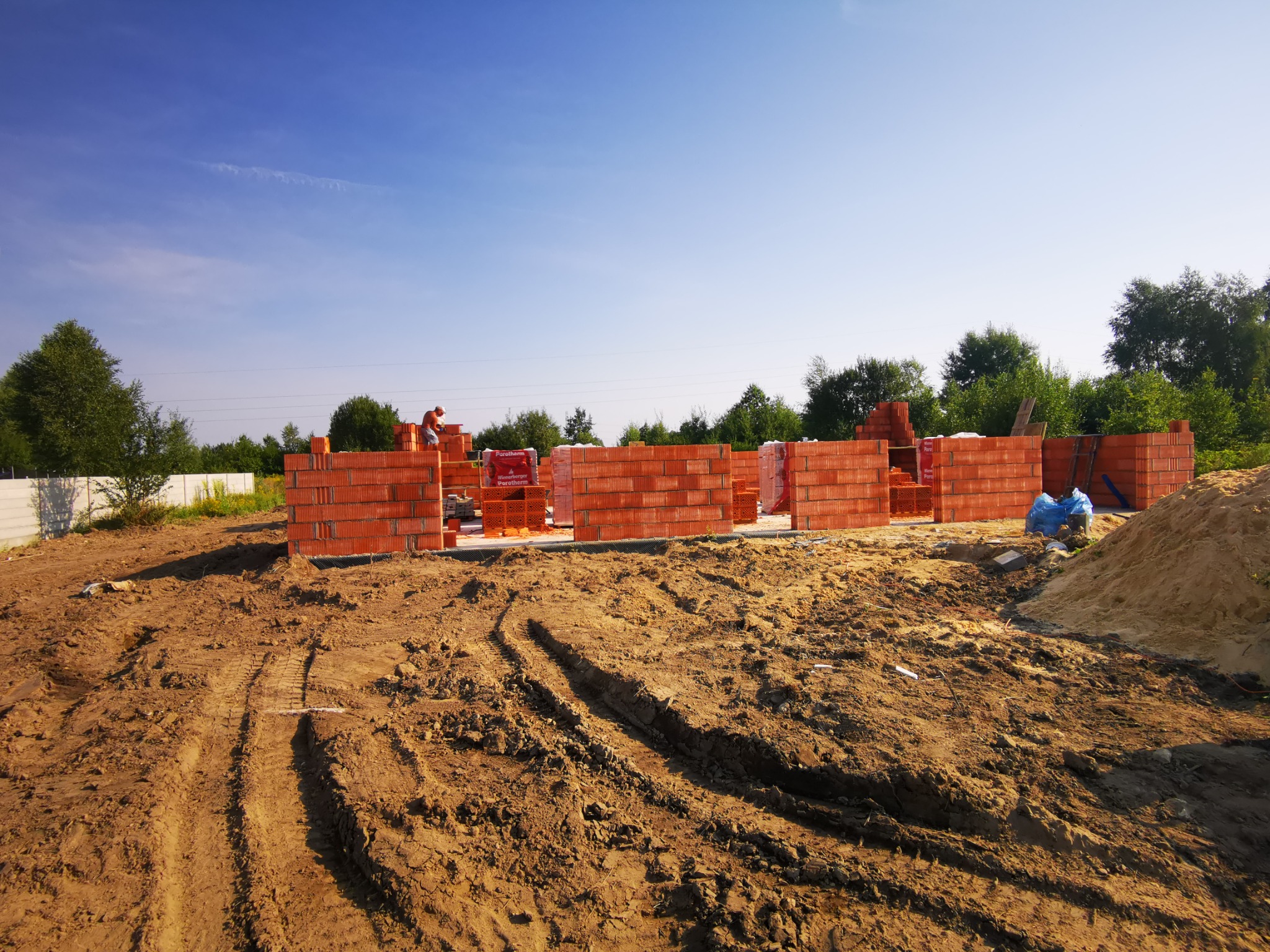 Wznoszenie ścian parterowego budynku z czerwonej ceramiki poryzowanej, widoczne ślady opon na placu budowy i pracownik w oddali.