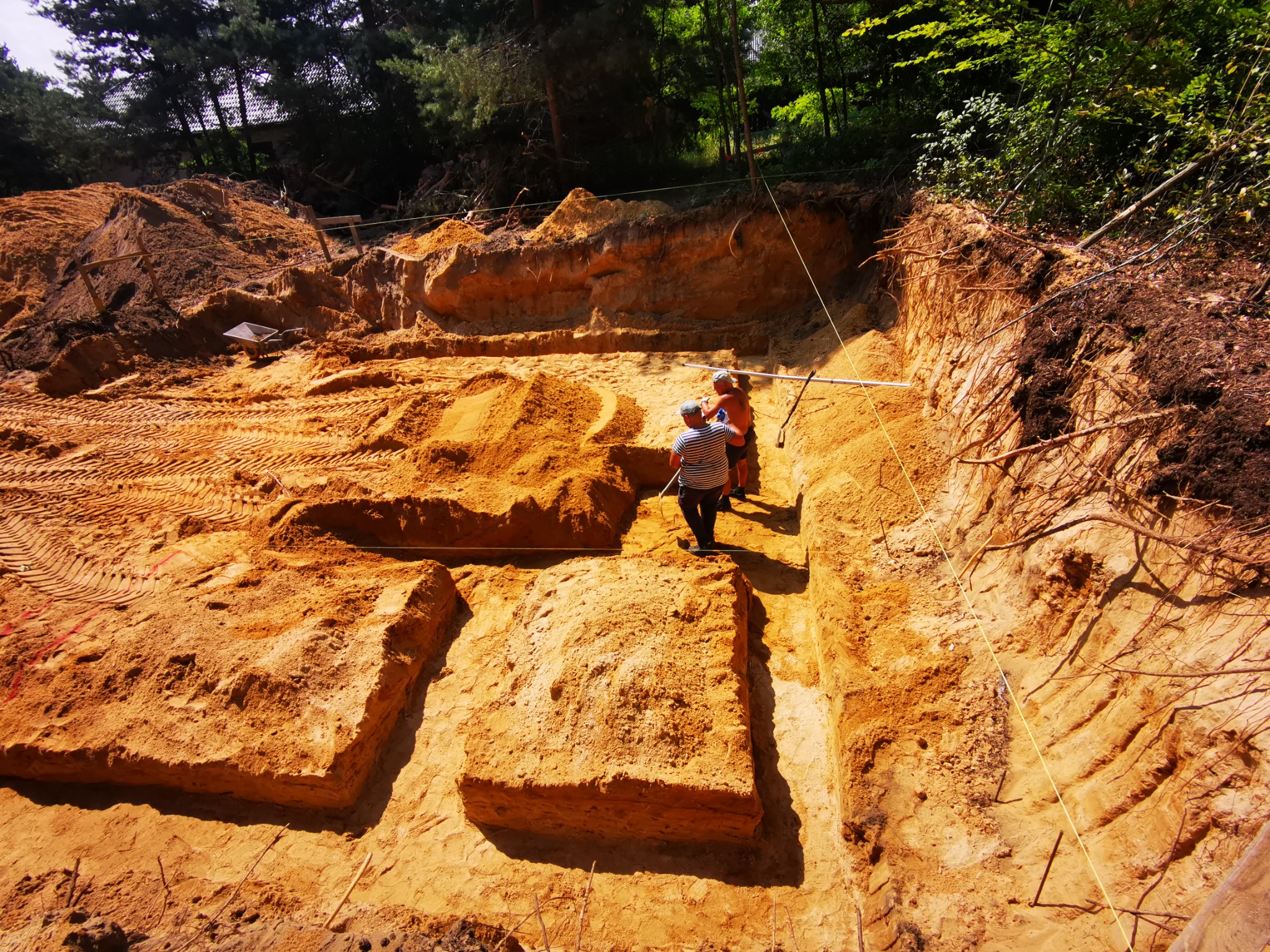 Wykop pod fundamenty z widocznymi wyznaczonymi obszarami, dwóch pracowników wewnątrz wykopu, teren piaszczysty, drzewa w tle.