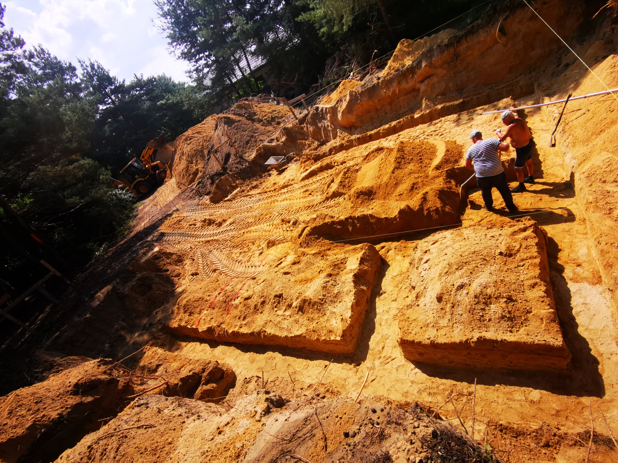 Wykop fundamentów w piaszczystym gruncie, z widocznymi śladami opon koparki i dwoma pracownikami wyrównującymi teren.