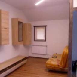 Pokój na poddaszu z drewnianą podłogą, meblami w odcieniach drewna i oświetleniem LED w suficie, widoczny fragment sofy i grzejnik pod oknem.