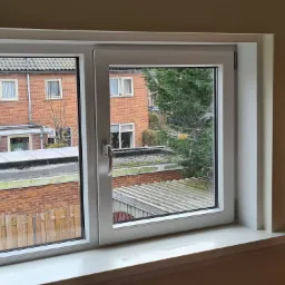 Białe, dwuskrzydłowe okno z widokiem na sąsiednie budynki z cegły i zielone drzewa, uchylone jedno skrzydło.