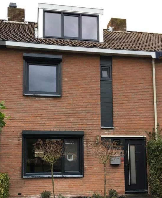 Dom z czerwonej cegły z szarymi oknami PCV i roletami zewnętrznymi w kolorze szarym, częściowo opuszczonymi na dwóch oknach, oraz szarymi drzwiami wejściowymi.