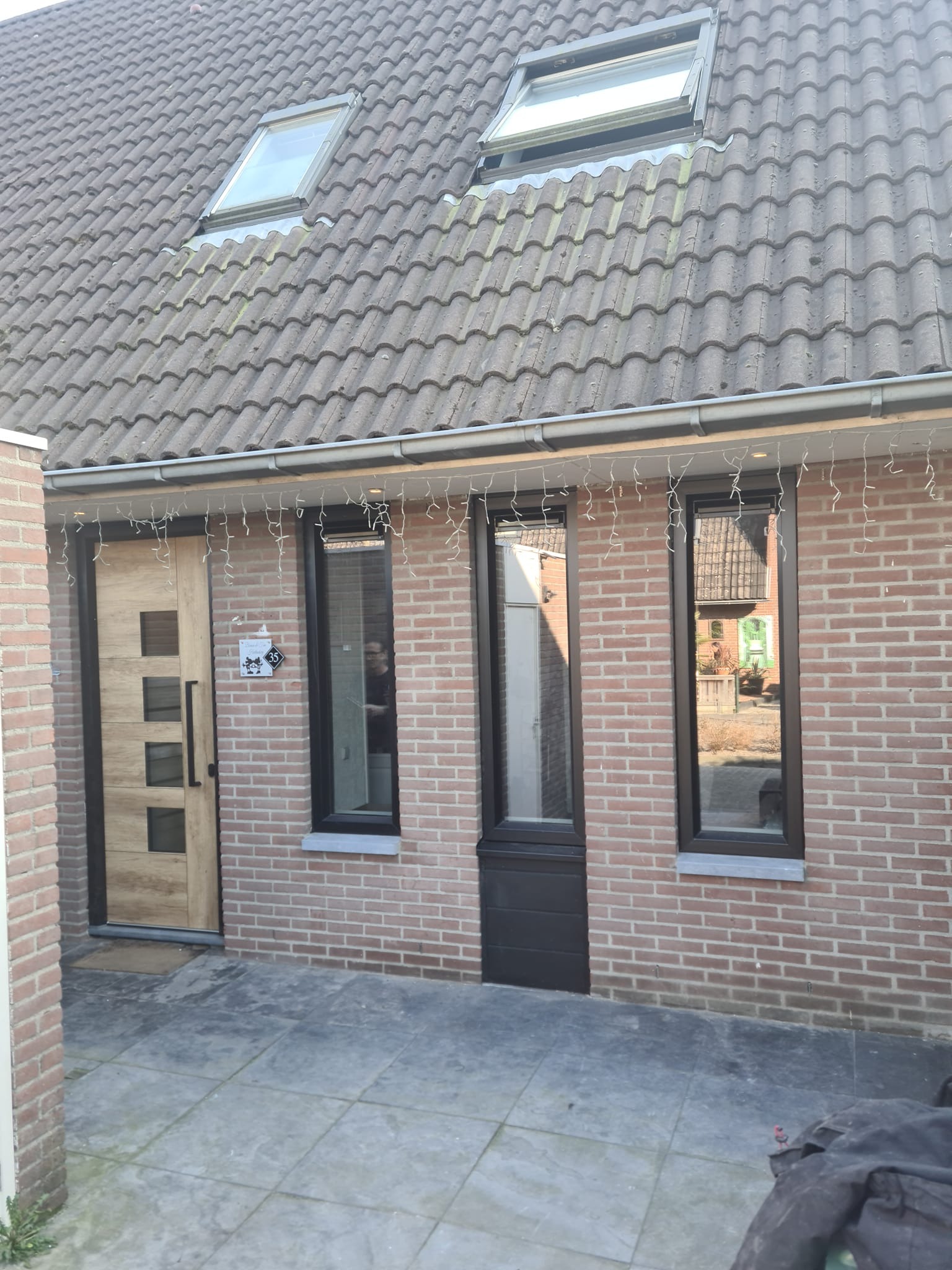 Dom z cegły z nowymi oknami z czarnymi ramami, drewnianymi drzwiami wejściowymi i dwoma oknami dachowymi, udekorowany świątecznymi lampkami.