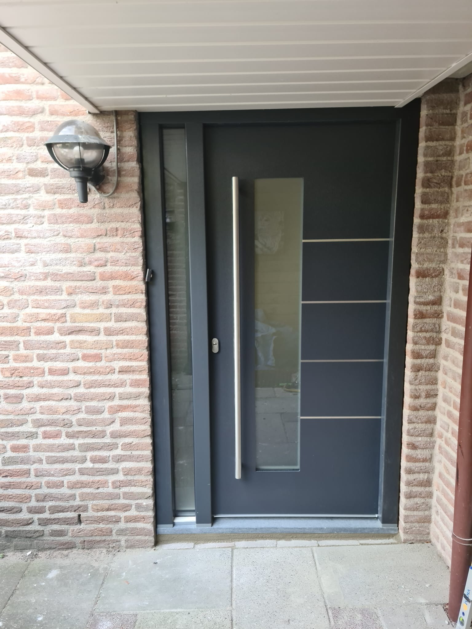 Nowoczesne, grafitowe drzwi wejściowe z przeszkleniem, osadzone w ceglanej ścianie, oświetlone lampą zewnętrzną.