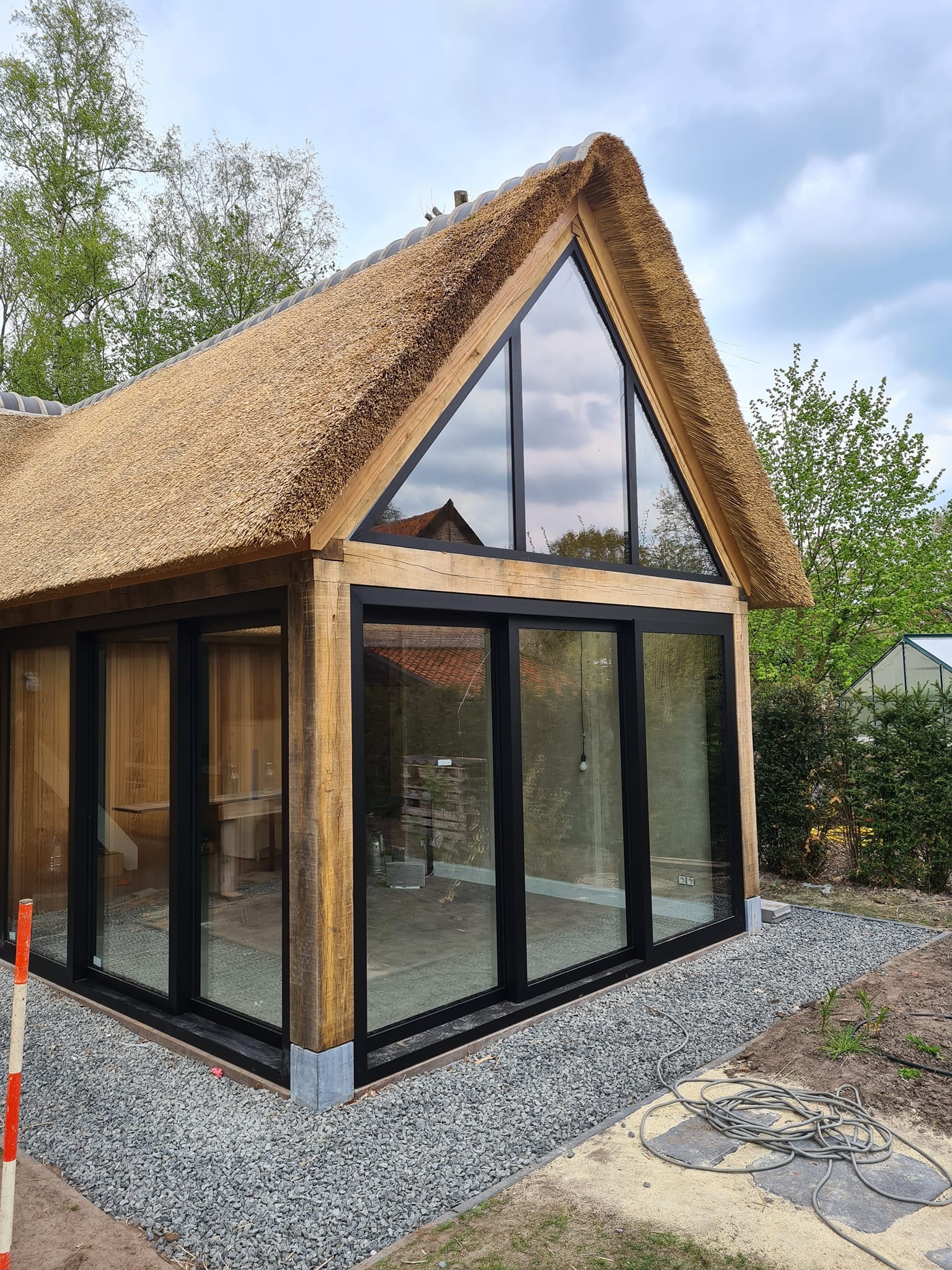 Nowoczesna oranżeria z dachem krytym strzechą i dużymi przeszkleniami z czarnymi ramami, otoczona żwirem i zielenią.