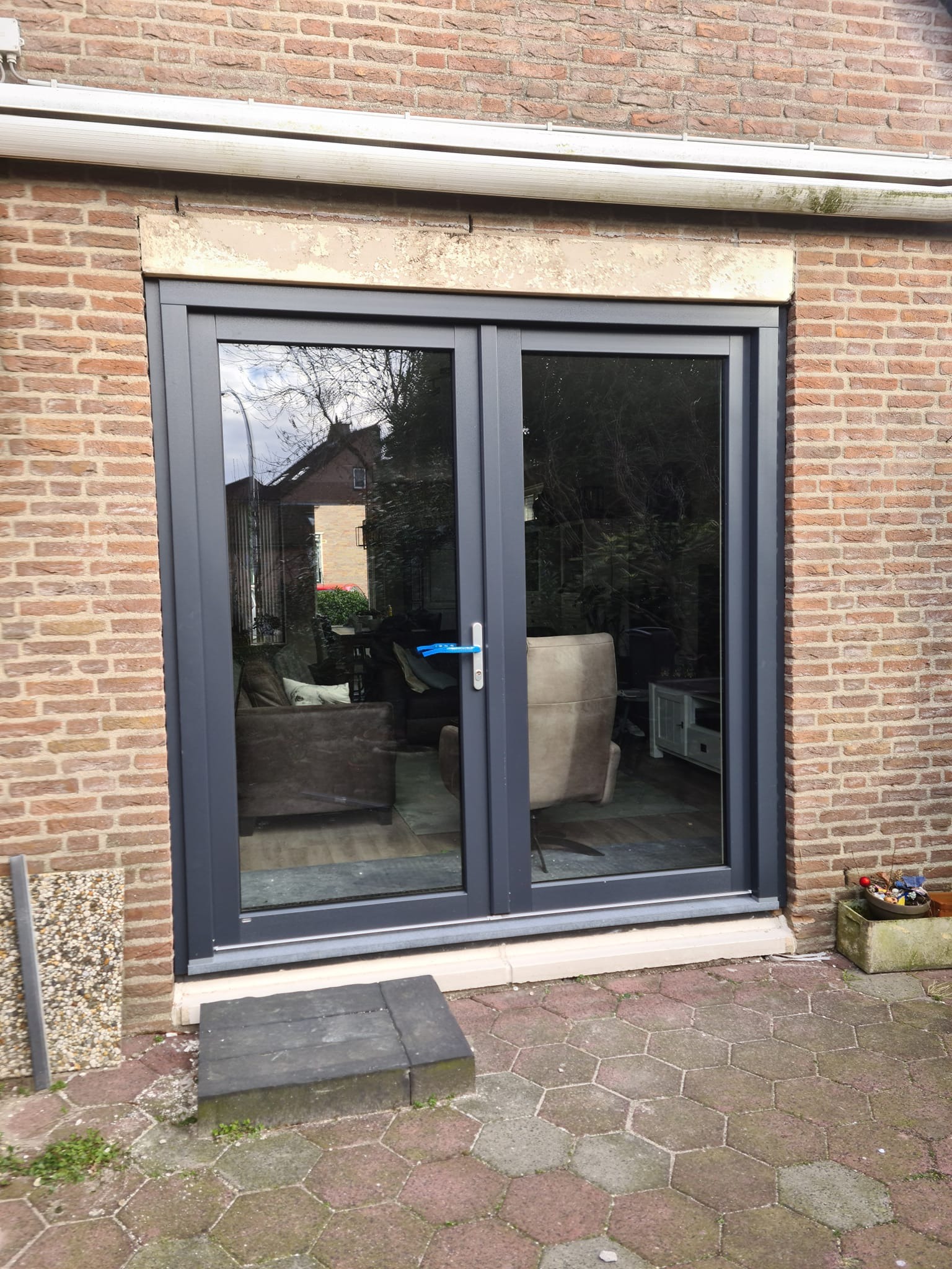 Szare dwuskrzydłowe drzwi balkonowe z widocznym odbiciem wnętrza domu, osadzone w murze z czerwonej cegły, z widocznym progiem i obrzeżem z kamienia.