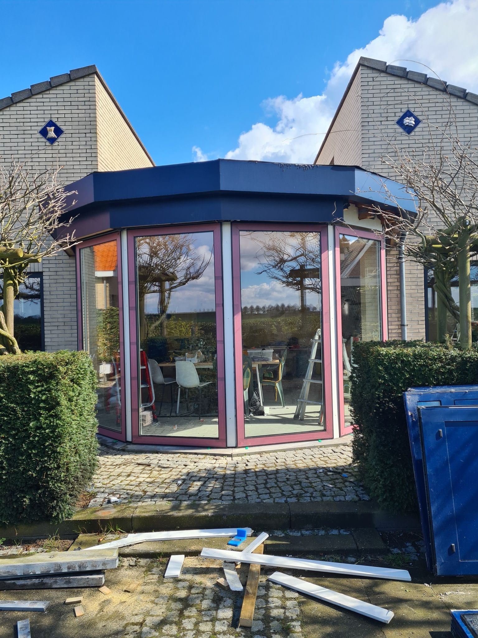 Ośmioboczna przeszklona weranda z różowymi ramami, widoczna z zewnątrz podczas montażu, z rozłożonymi elementami konstrukcyjnymi na ziemi i drabiną wewnątrz.