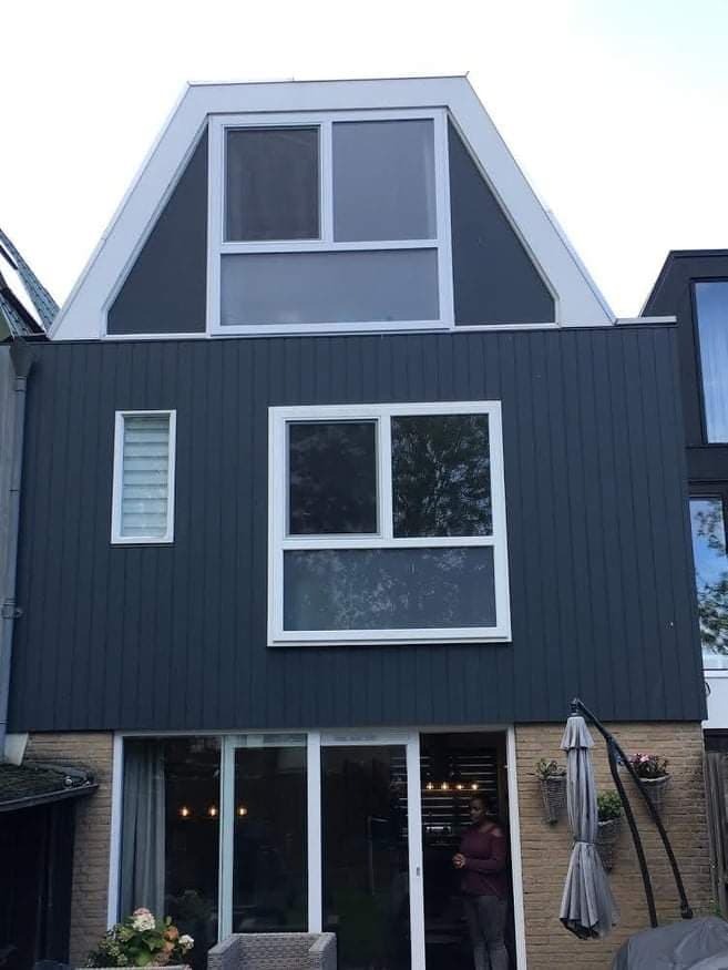 Dwukondygnacyjny dom z szarą elewacją i białymi ramami okiennymi, widoczne okno PCV na poddaszu, duże okno na parterze i przesuwne drzwi balkonowe, z boku mur z cegły