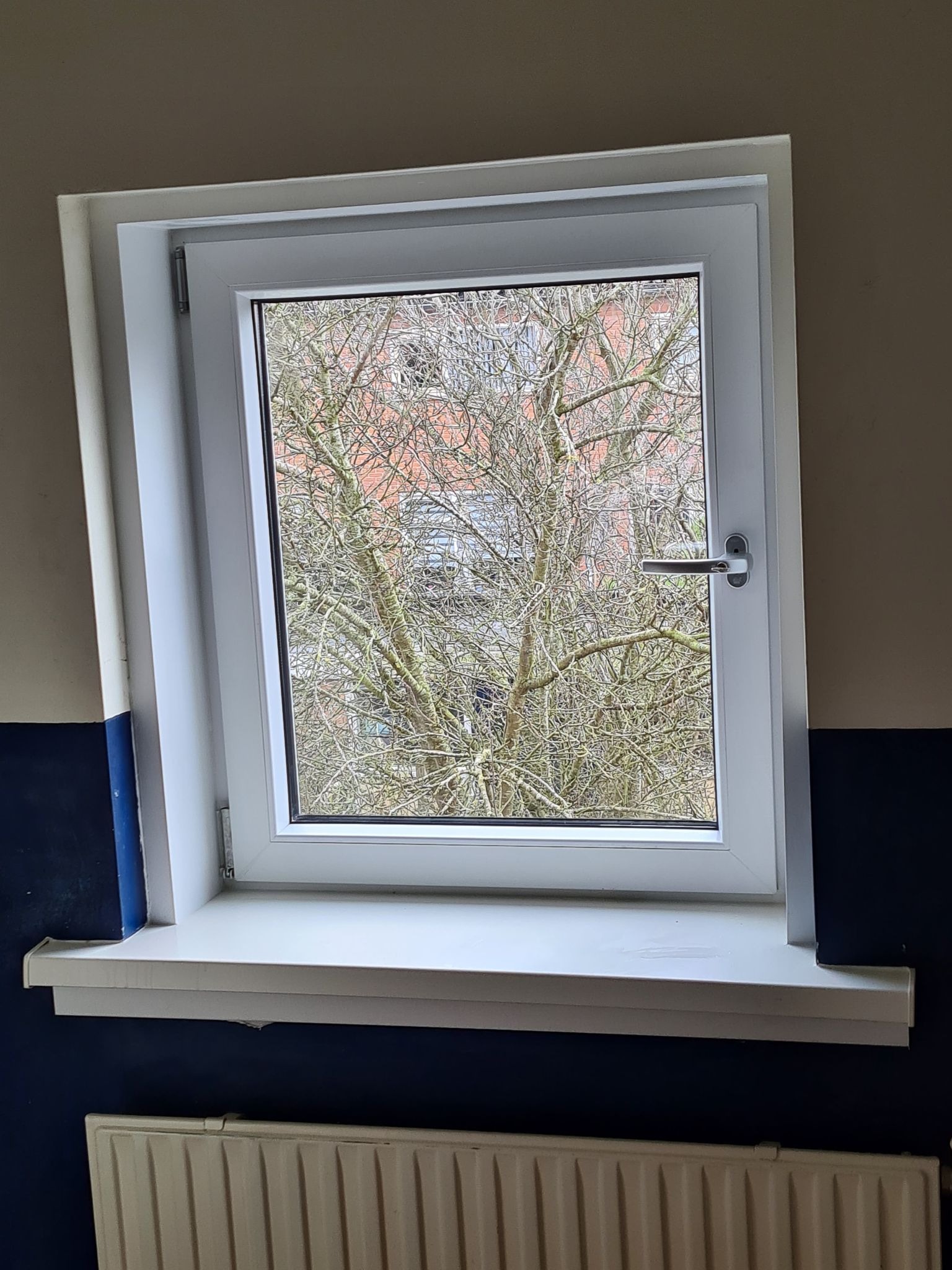 Białe okno PCV z widokiem na drzewo bez liści w Dordrecht, z widocznym grzejnikiem pod parapetem i dwukolorową ścianą.