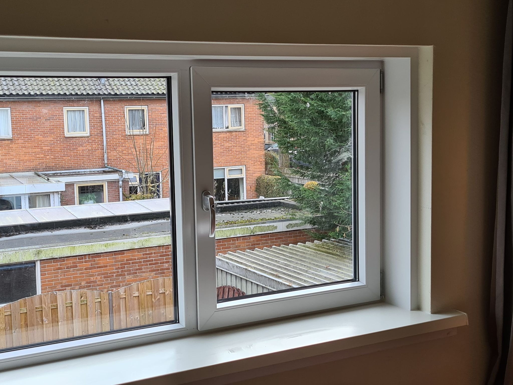 Białe, dwuskrzydłowe okno z widokiem na sąsiednie budynki z cegły i zielone drzewa, uchylone jedno skrzydło.