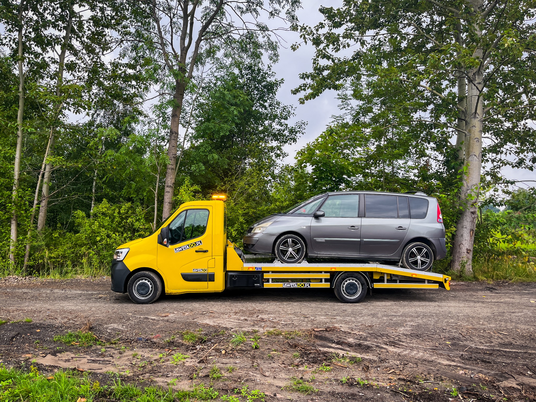 Żółta laweta z szarym vanem na platformie, sfotografowana w Dąbrowie Górniczej na tle drzew, z widocznym napisem 'LAWETA DO.PL' na pojeździe.