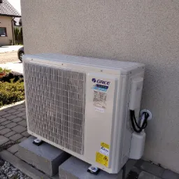 Zewnętrzna jednostka klimatyzacji Gree Inverter zamontowana na betonowych bloczkach przy elewacji budynku z widocznymi przewodami i fragmentem ogrodu.