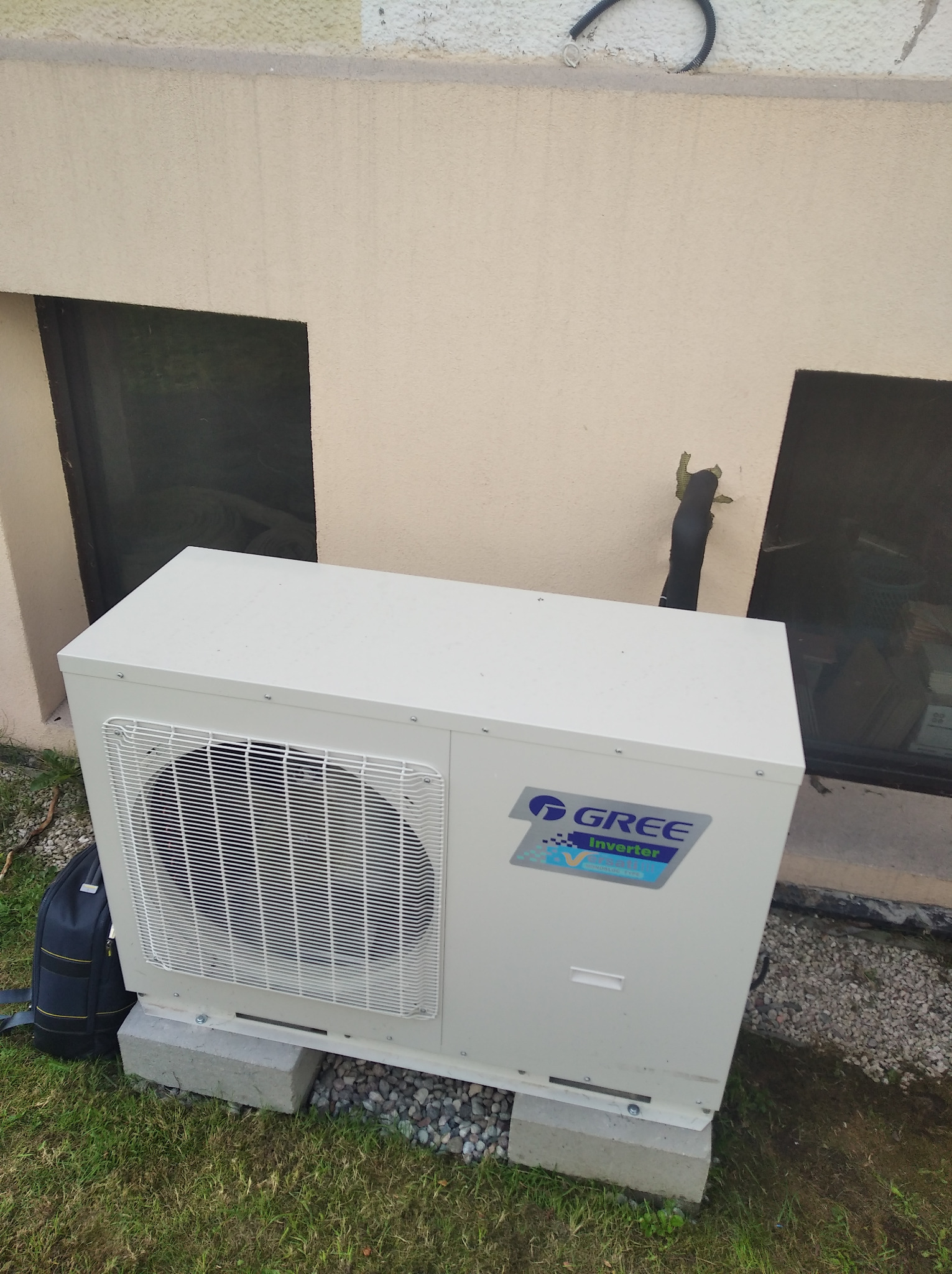 Zewnętrzna jednostka klimatyzacji Gree Inverter zamontowana na betonowych bloczkach przy ścianie budynku w Gdyni, widoczny fragment trawnika i okno piwniczne.