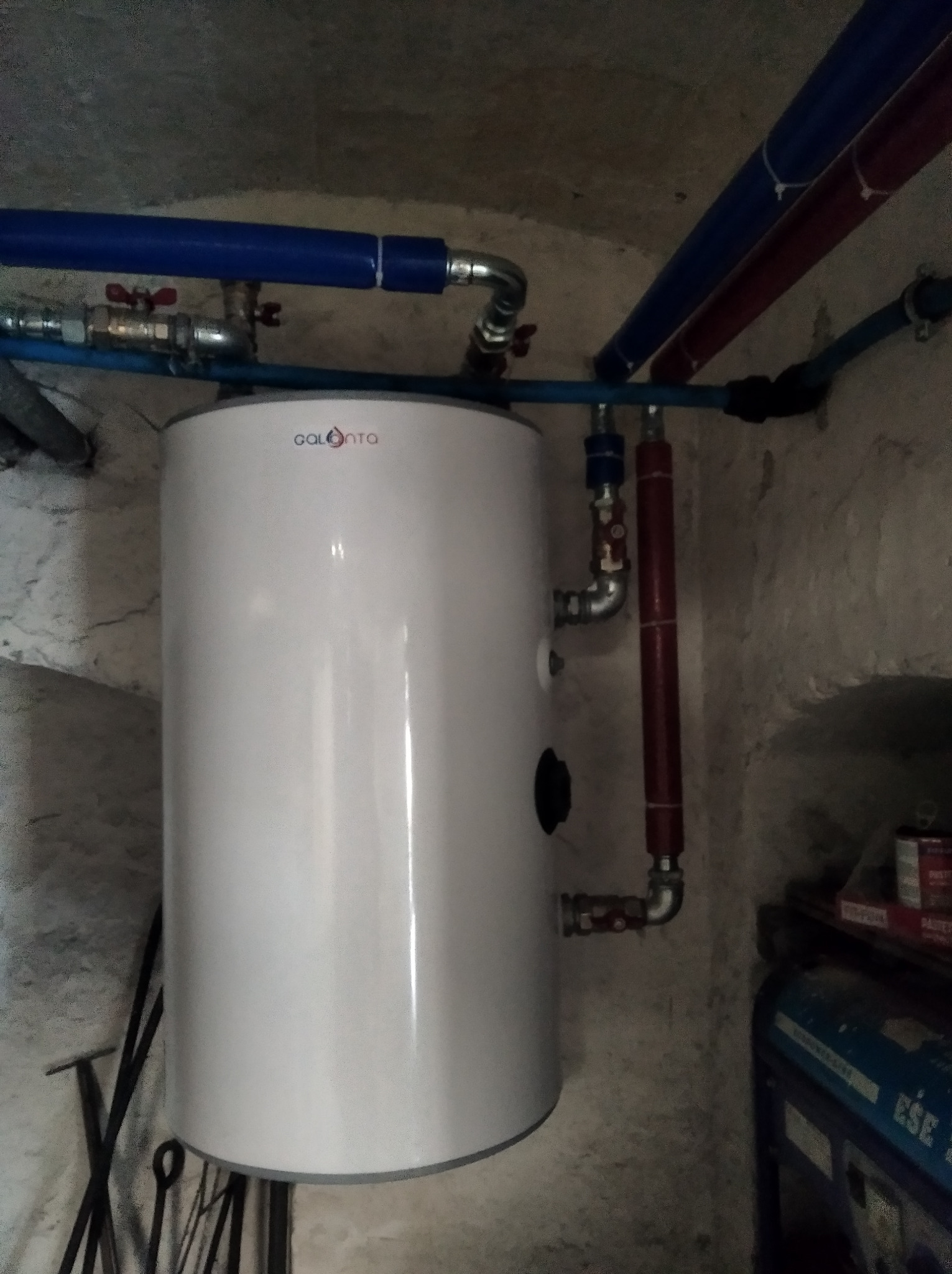 Bojler Galmet z niebieskimi i czerwonymi rurami w surowym pomieszczeniu z szarą ścianą i narzędziami w tle.