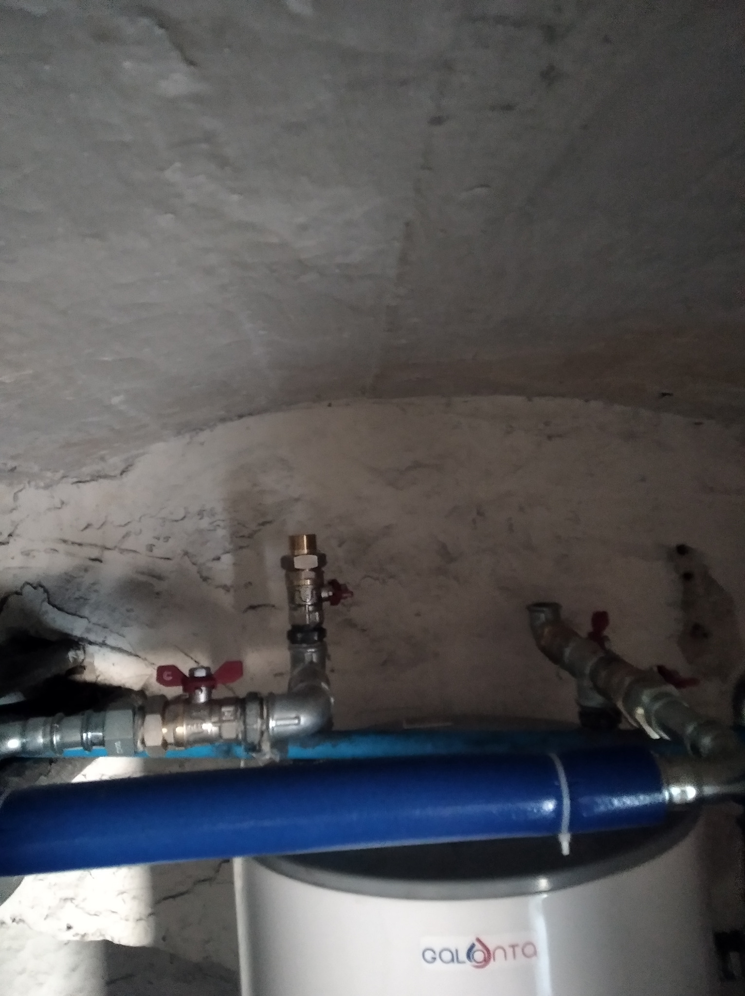 Instalacja hydrauliczna z zaworami i niebieską rurą nad bojlerem marki Galmet, widoczne rury miedziane i białe ściany w tle.