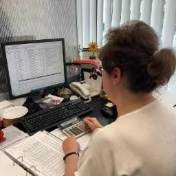 Kobieta przy biurku, odwrócona tyłem, używa kalkulatora nad otwartym segregatorze z tabelami, monitor komputera wyświetla arkusz kalkulacyjny, na biurku telefon i inne akcesoria biurowe.