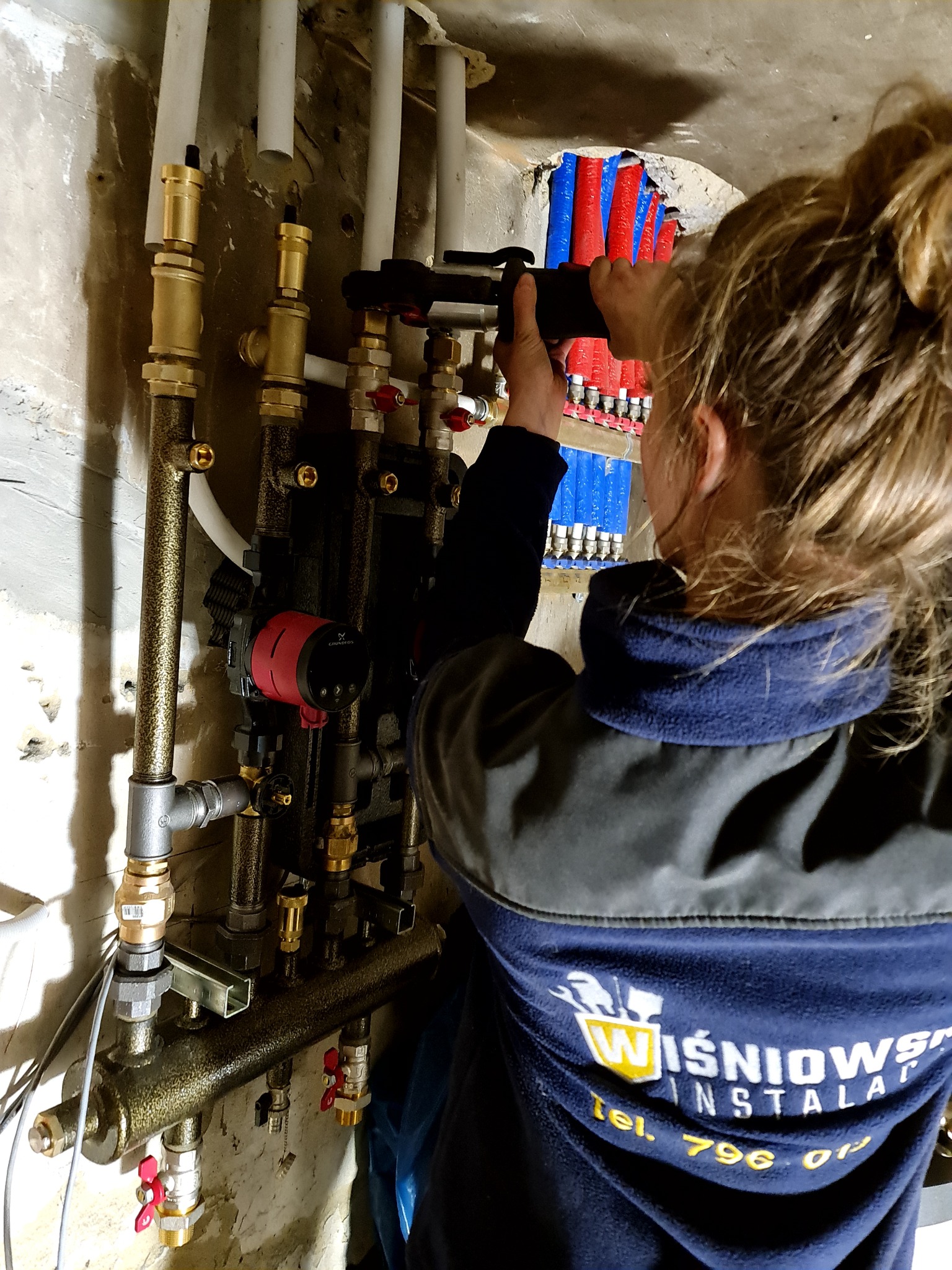 Instalator zaciska podejścia do rozdzielacza ogrzewania podłogowego; widoczny rozdzielacz z rurami w kolorze czerwonym i niebieskim, oraz pompa obiegowa Grundfos w kolorze czerwono-czarnym...