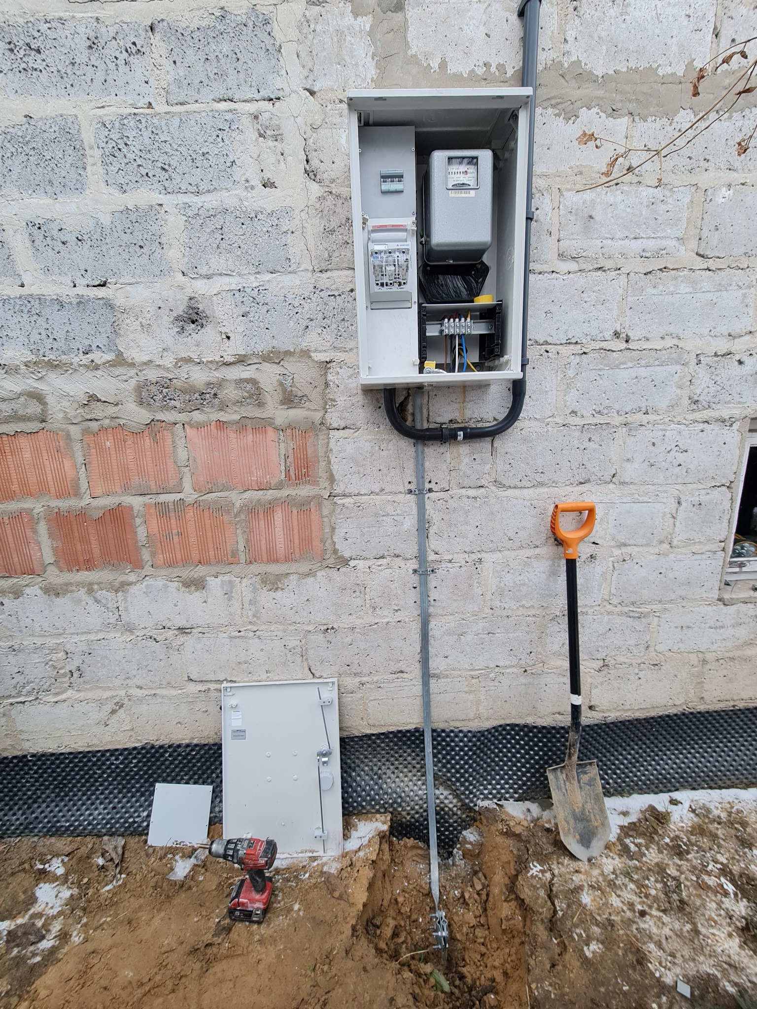 Montaż skrzynki z licznikiem energii elektrycznej na ścianie z bloczków, widoczne uziemienie pionowe, szpadel i wiertarka akumulatorowa.
