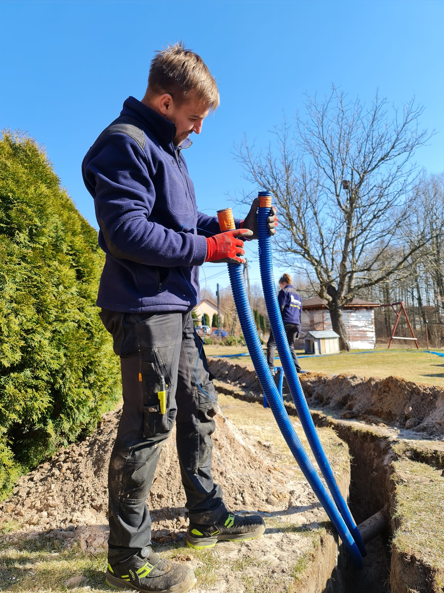 Instalator w roboczych ubraniach, w trakcie rozprowadzania dwóch niebieskich rur karbowanych w wykopie ziemnym na posesji, widoczna druga osoba i elementy wyposażenia ogrodu w tle.