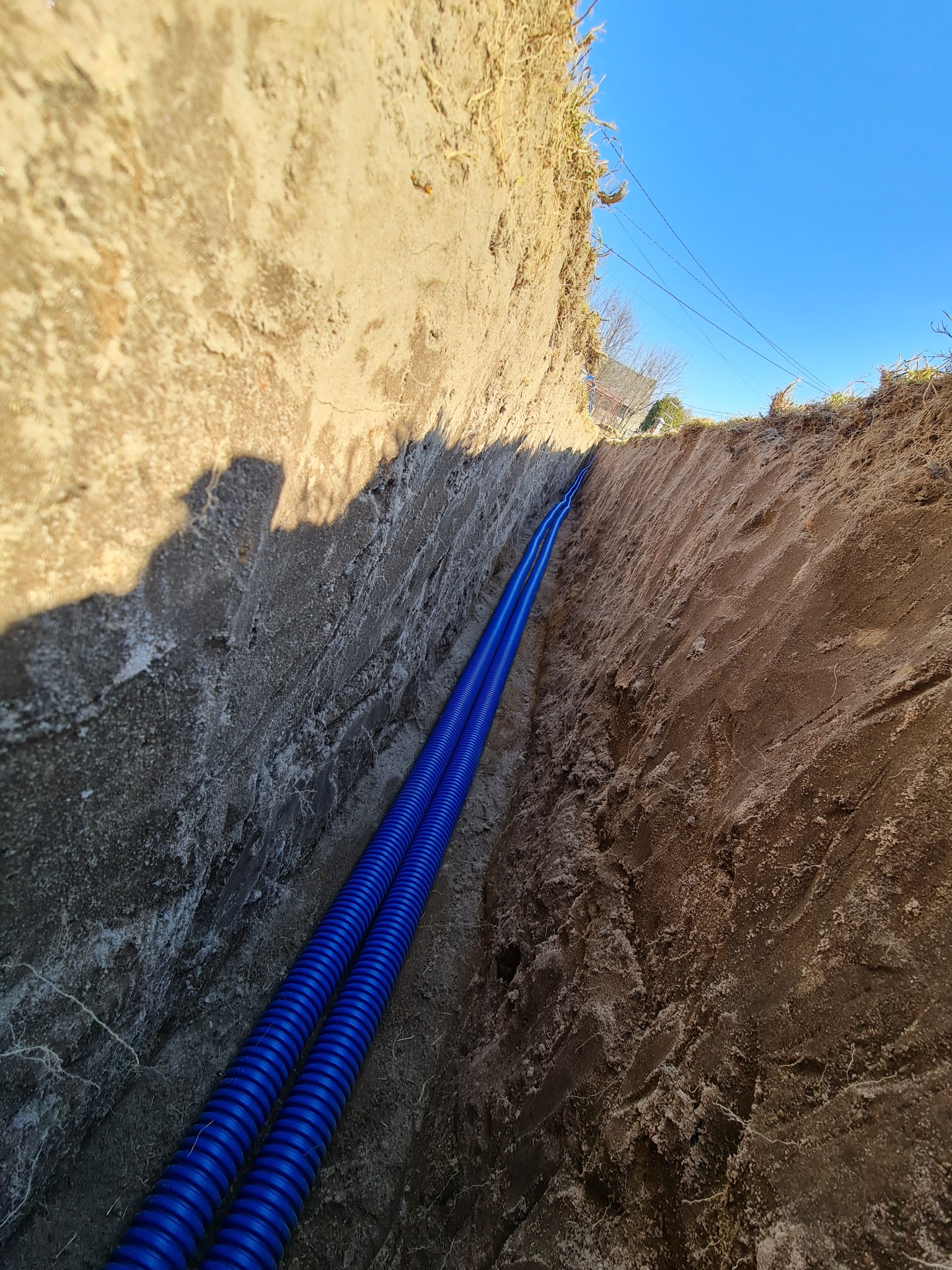 Dwa niebieskie karbowane przewody ułożone równolegle w wykopie o ziemnych ścianach. Widoczny cień osoby robiącej zdjęcie.