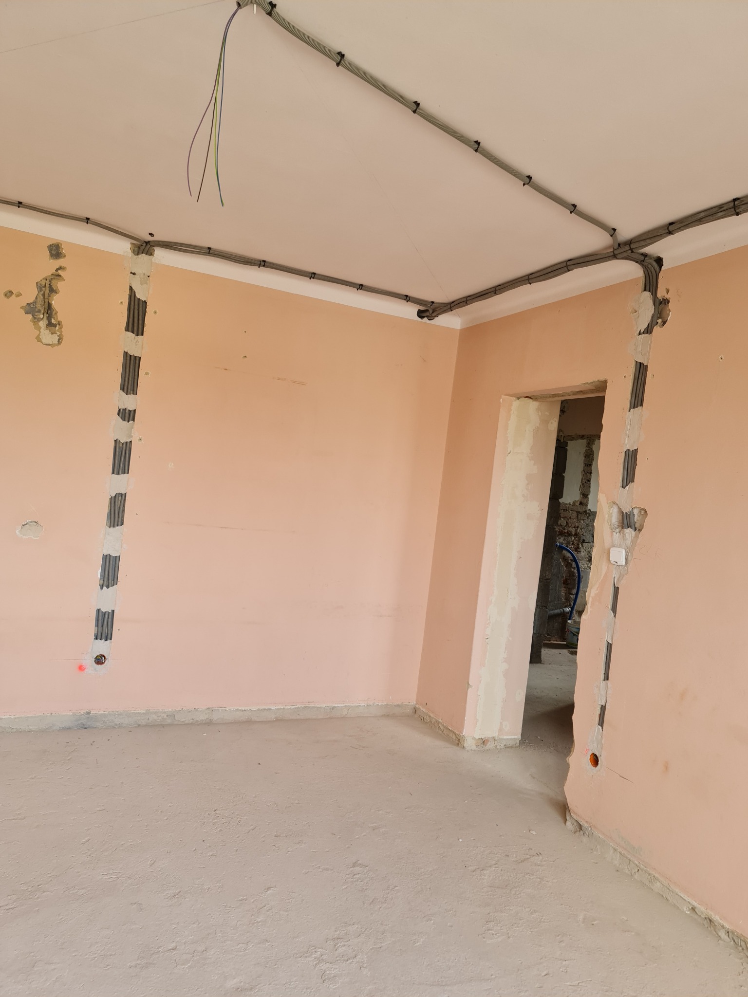 Wnętrze w trakcie remontu, widoczne kable elektryczne w peszlach prowadzone po ścianach i suficie, przygotowane podtynkowo gniazdka, ściany w różowym kolorze przed położeniem gładzi.