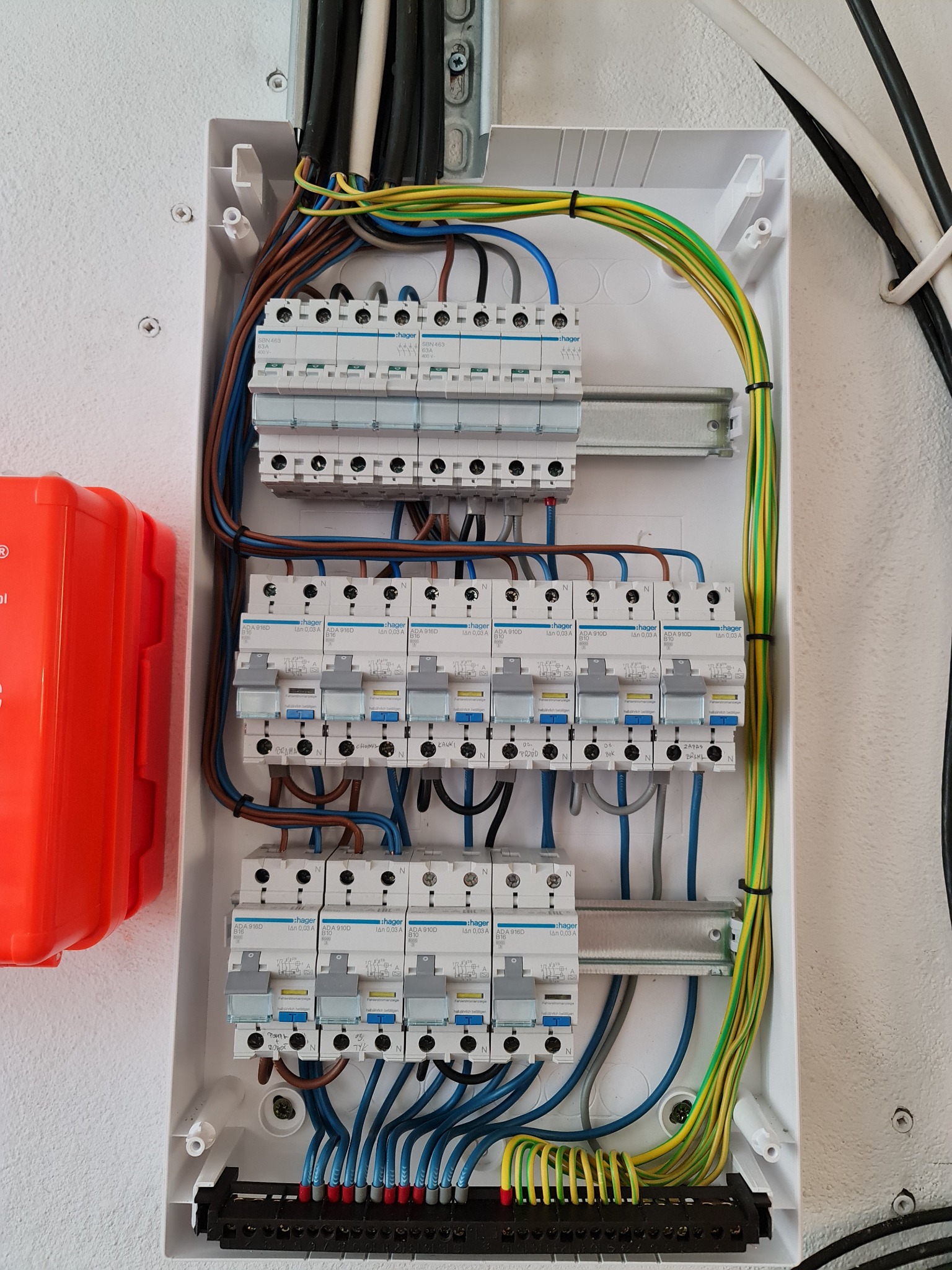 Otwarta skrzynka rozdzielcza z bezpiecznikami Hager, widoczne przewody w kolorach brązowym, niebieskim i żółto-zielonym, oraz pomarańczowa obudowa zabezpieczająca z boku.