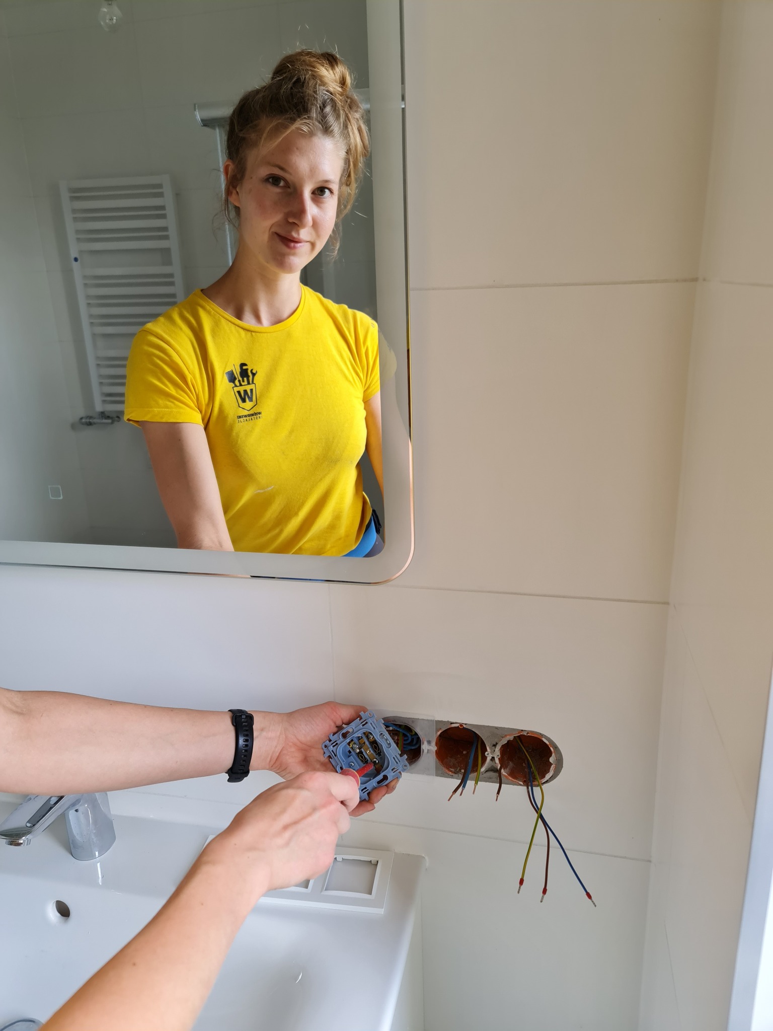 Elektryk w żółtej koszulce z logo firmy montuje gniazdko elektryczne w łazience; widoczne przewody w ścianie i odbicie kobiety w lustrze.