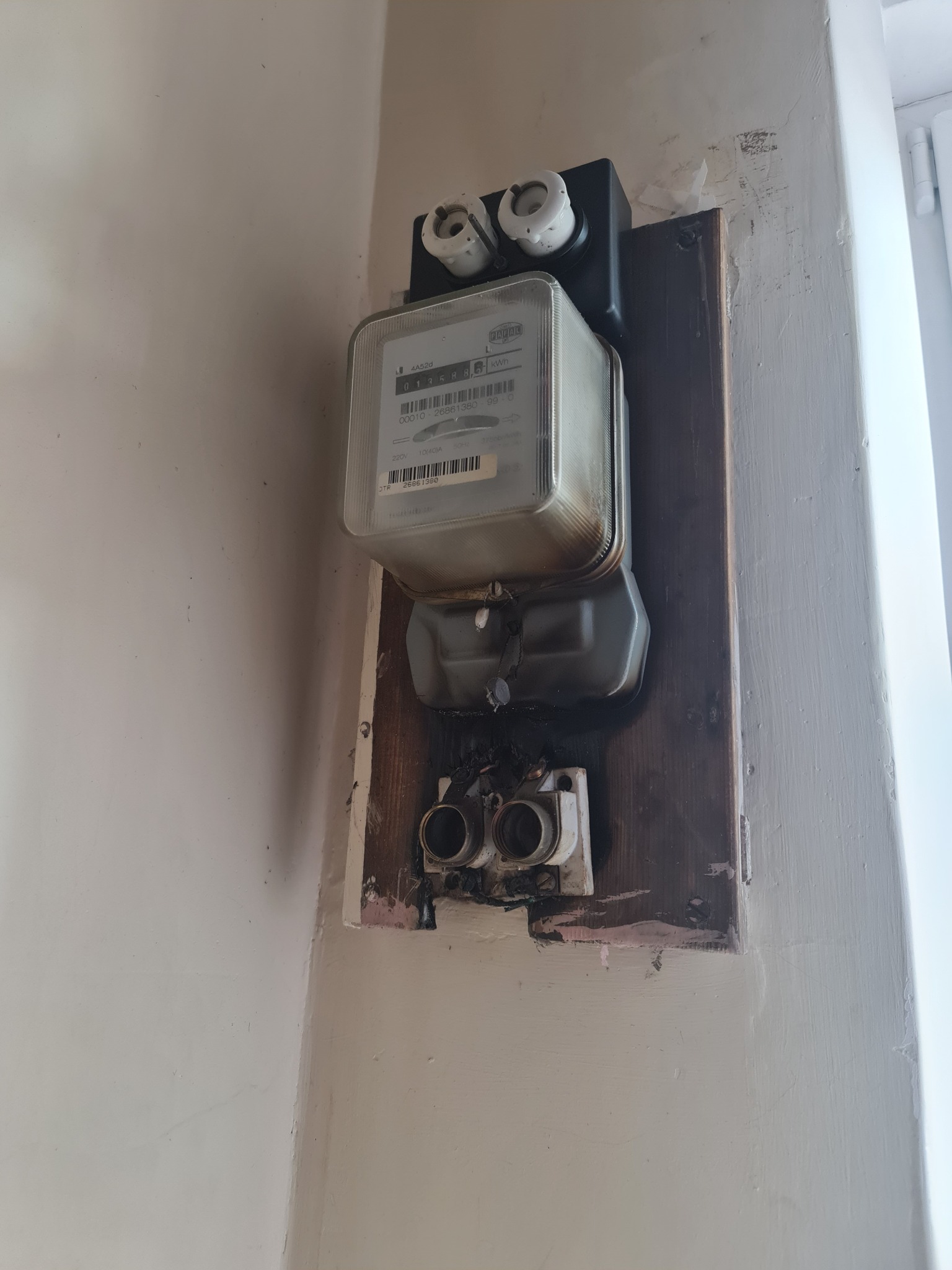 Stary, zużyty licznik elektryczny zamontowany na drewnianej płycie przy ścianie z widocznymi śladami uszkodzeń i napraw.