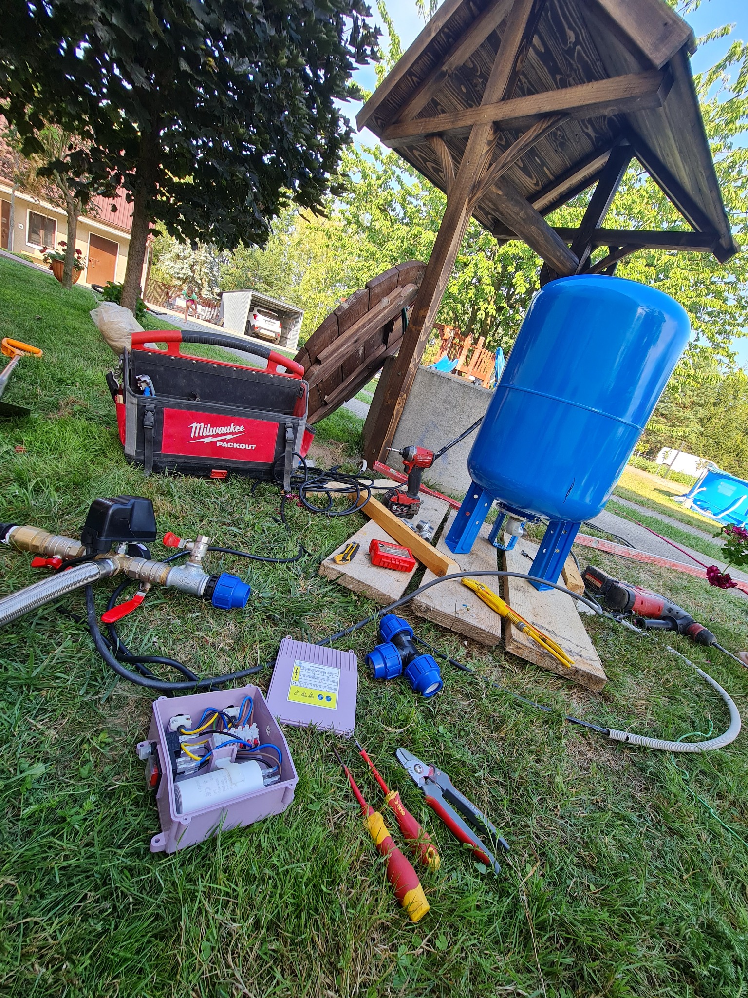 Niebieski zbiornik hydroforowy na trawie, obok skrzynka narzędziowa Milwaukee, otwarte pudełko elektryczne z kondensatorem, narzędzia hydrauliczne i elektryczne, drewniana konstrukcja zadaszenia...