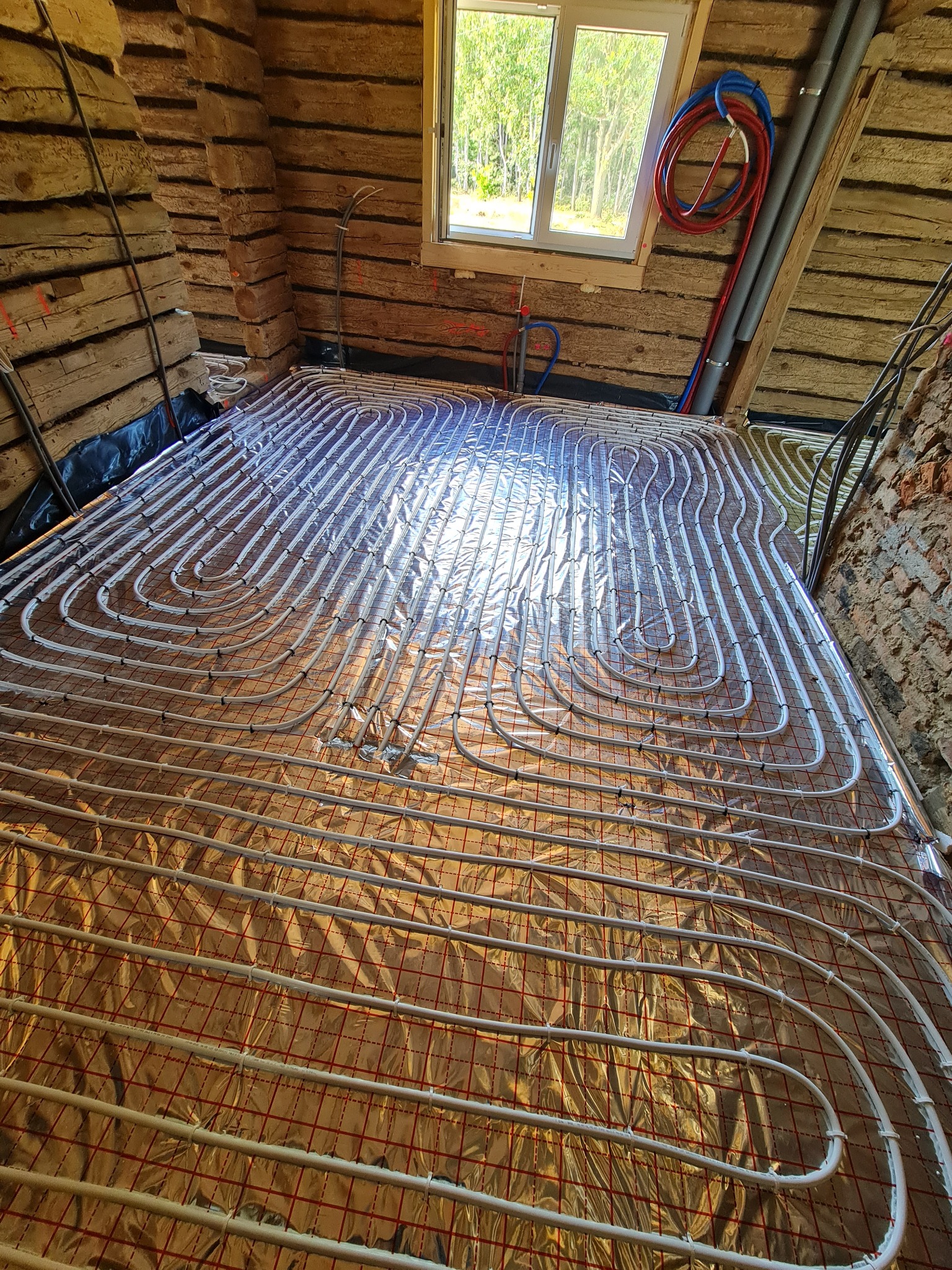 Instalacja ogrzewania podłogowego z rurkami na folii izolacyjnej w drewnianym domu z oknem i widokiem na drzewa.