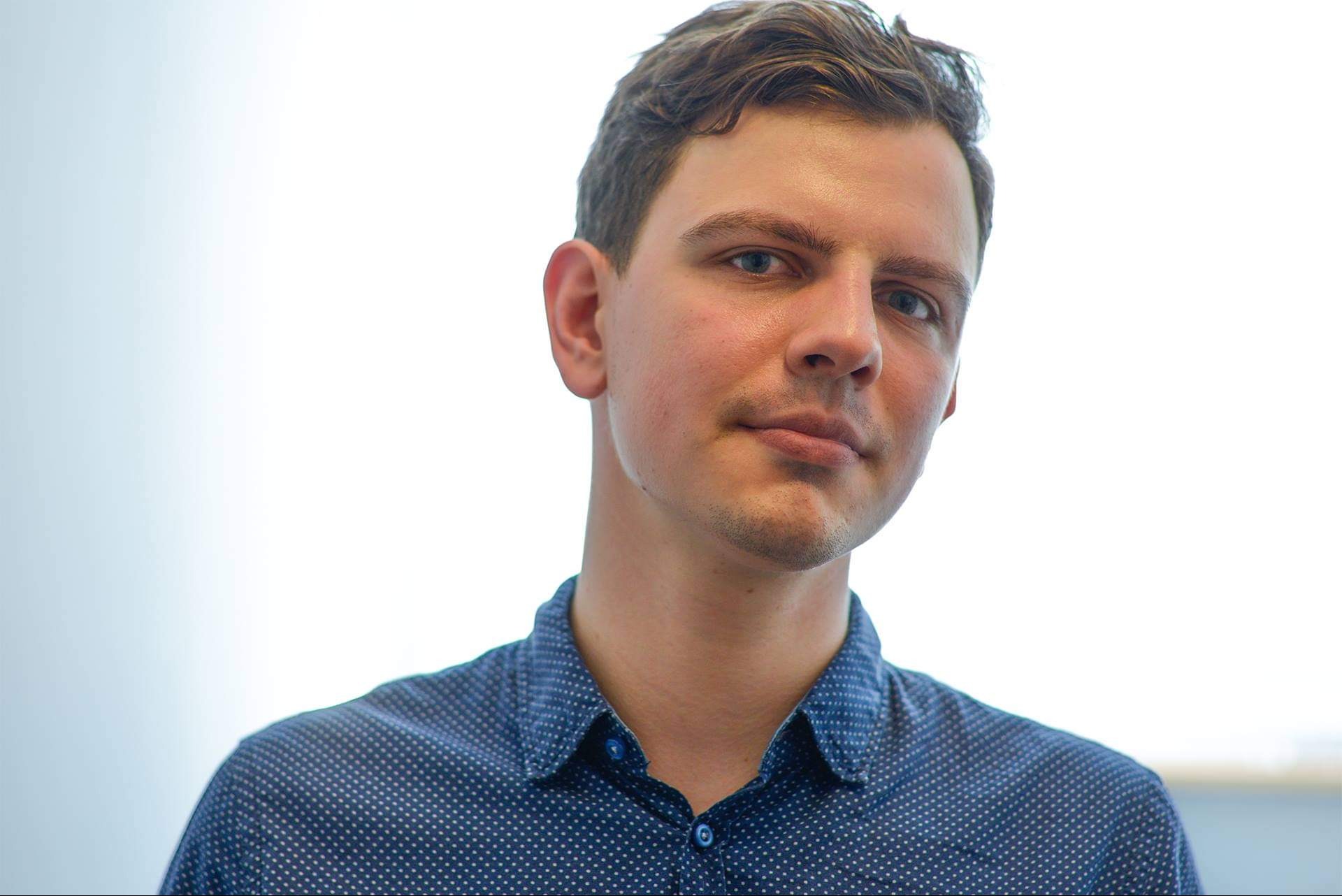 Portret młodego mężczyzny w niebieskiej koszuli w kropki, z krótkimi brązowymi włosami i niebieskimi oczami, na jasnym tle.