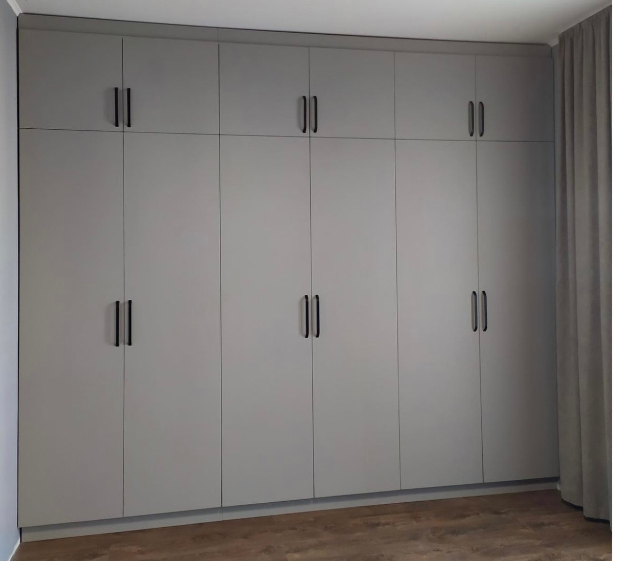 Duża, szara szafa wnękowa z czarnymi, pionowymi uchwytami, zajmująca całą ścianę, z zasłoną po prawej stronie i podłogą imitującą drewno.