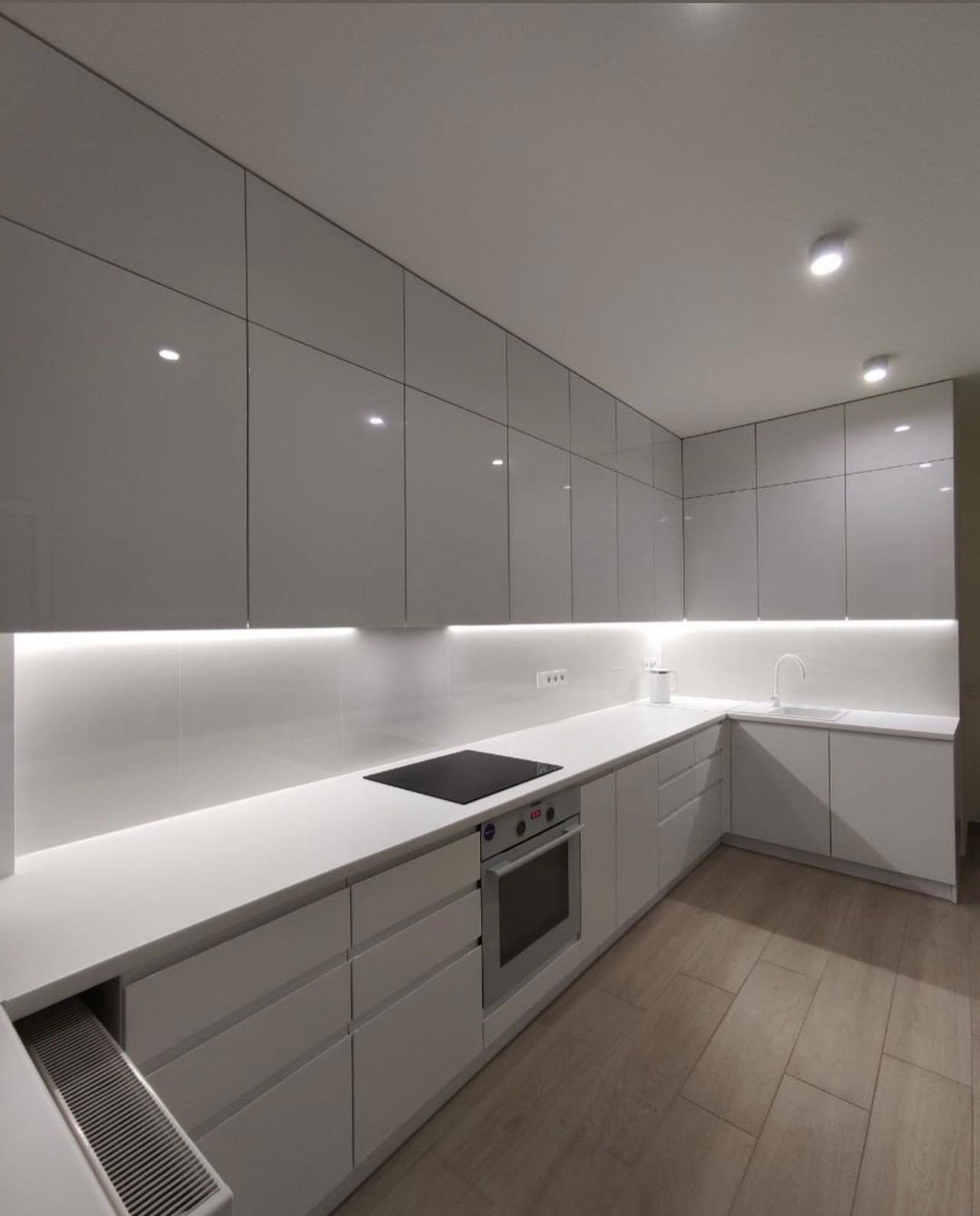 Nowoczesna, minimalistyczna kuchnia z białymi, lakierowanymi frontami i podświetleniem LED pod szafkami wiszącymi, widok na płytę indukcyjną i piekarnik w zabudowie.