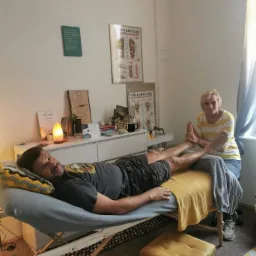 Mężczyzna leży na kozetce podczas masażu stóp wykonywanego przez kobietę w żółto-białej bluzce; w tle dyplom, lampka solna i plansze refleksologii stóp i dłoni.